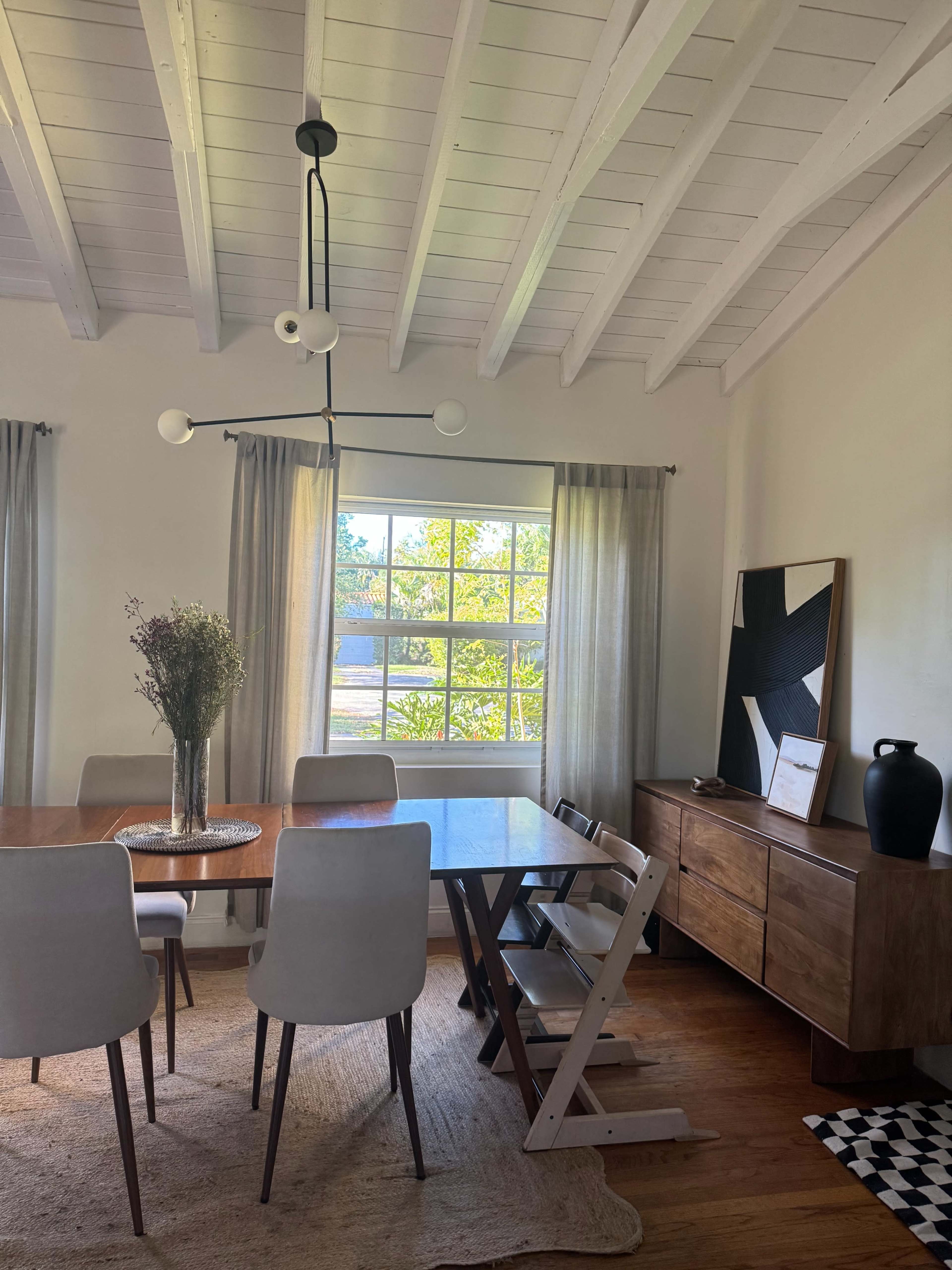 A dining room features a wooden table surrounded by chairs, a large window with natural light, and a sideboard with decorative items.
