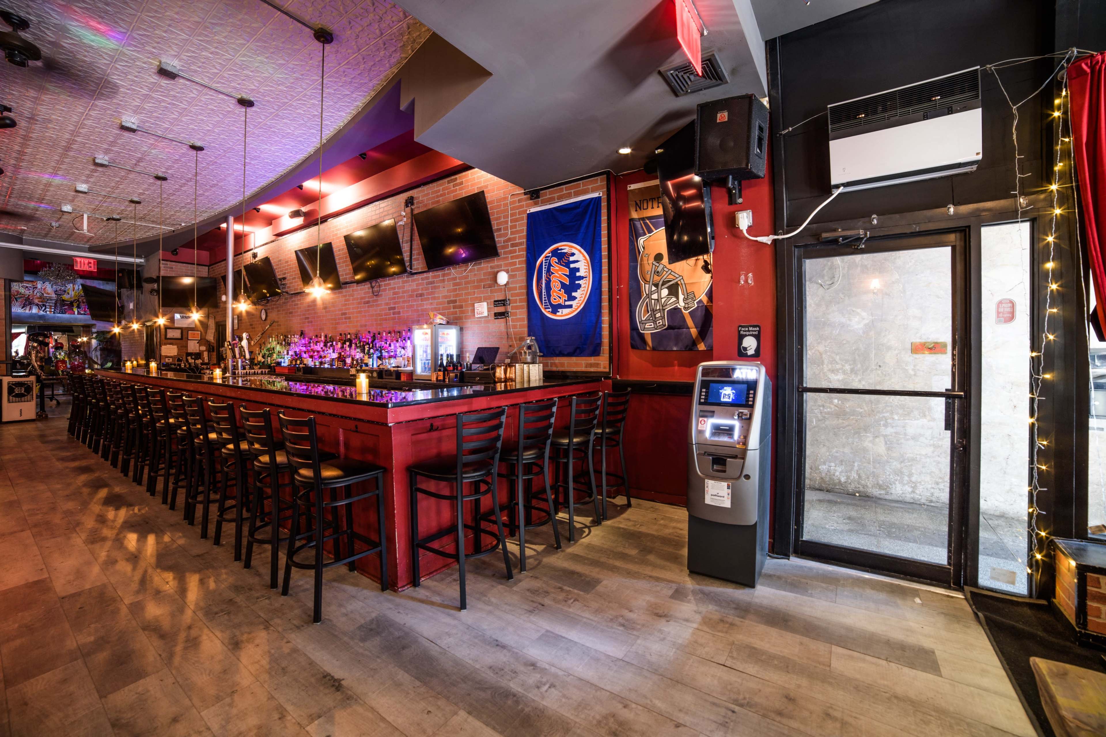 The image shows a bar area with a long counter, several bar stools, and wall-mounted televisions, illuminated by ambient lighting.