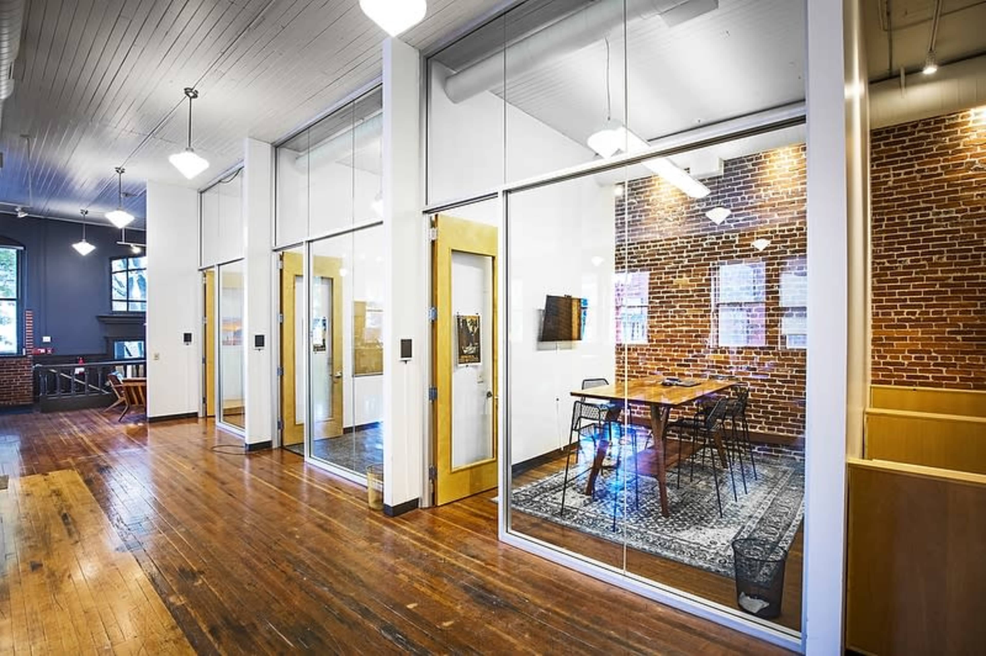 A modern office space with glass-enclosed meeting rooms, exposed brick walls, and a wooden floor.