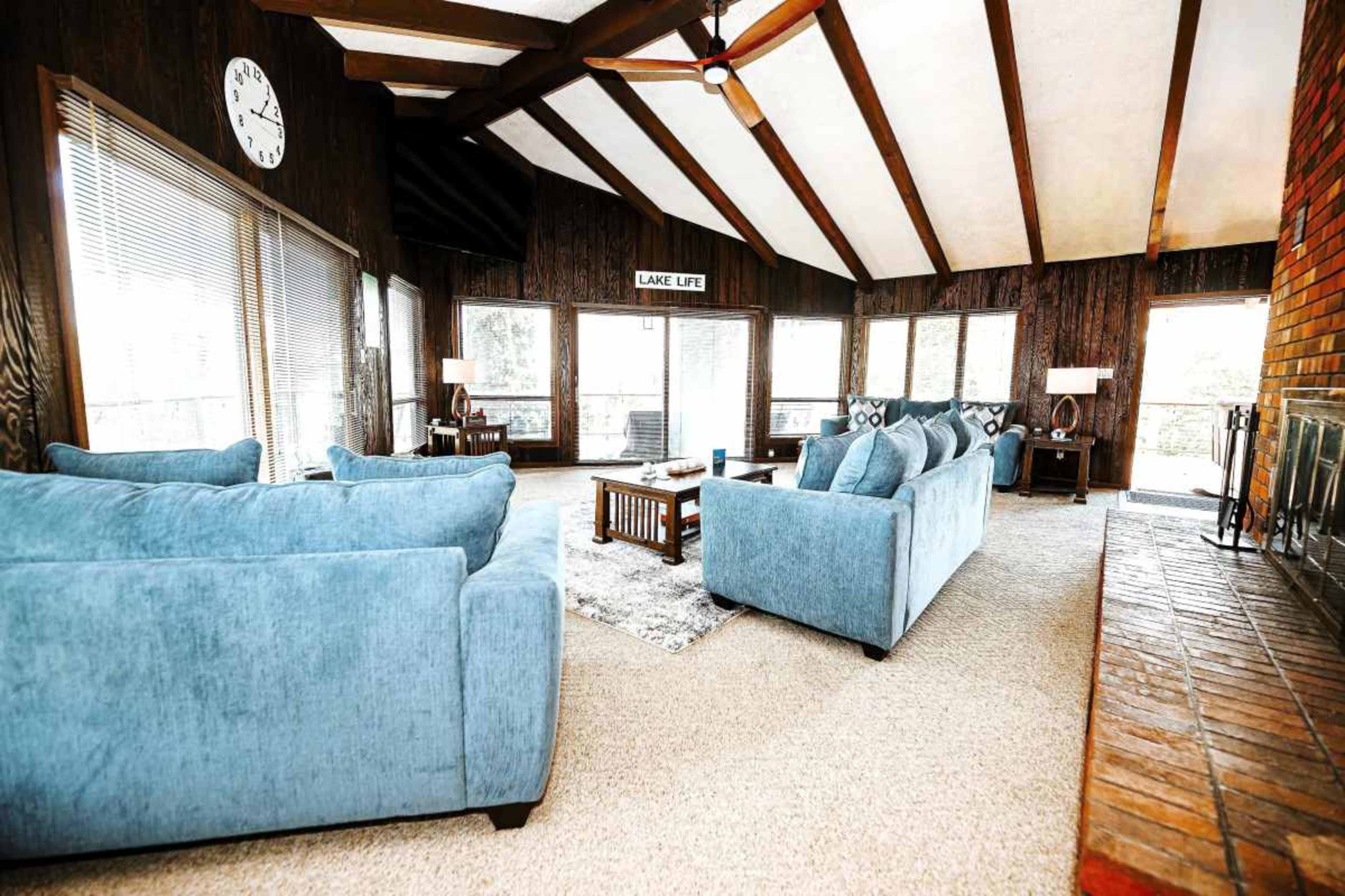 The image shows a spacious living room with wooden beams, large windows, blue sofas, and a brick fireplace.