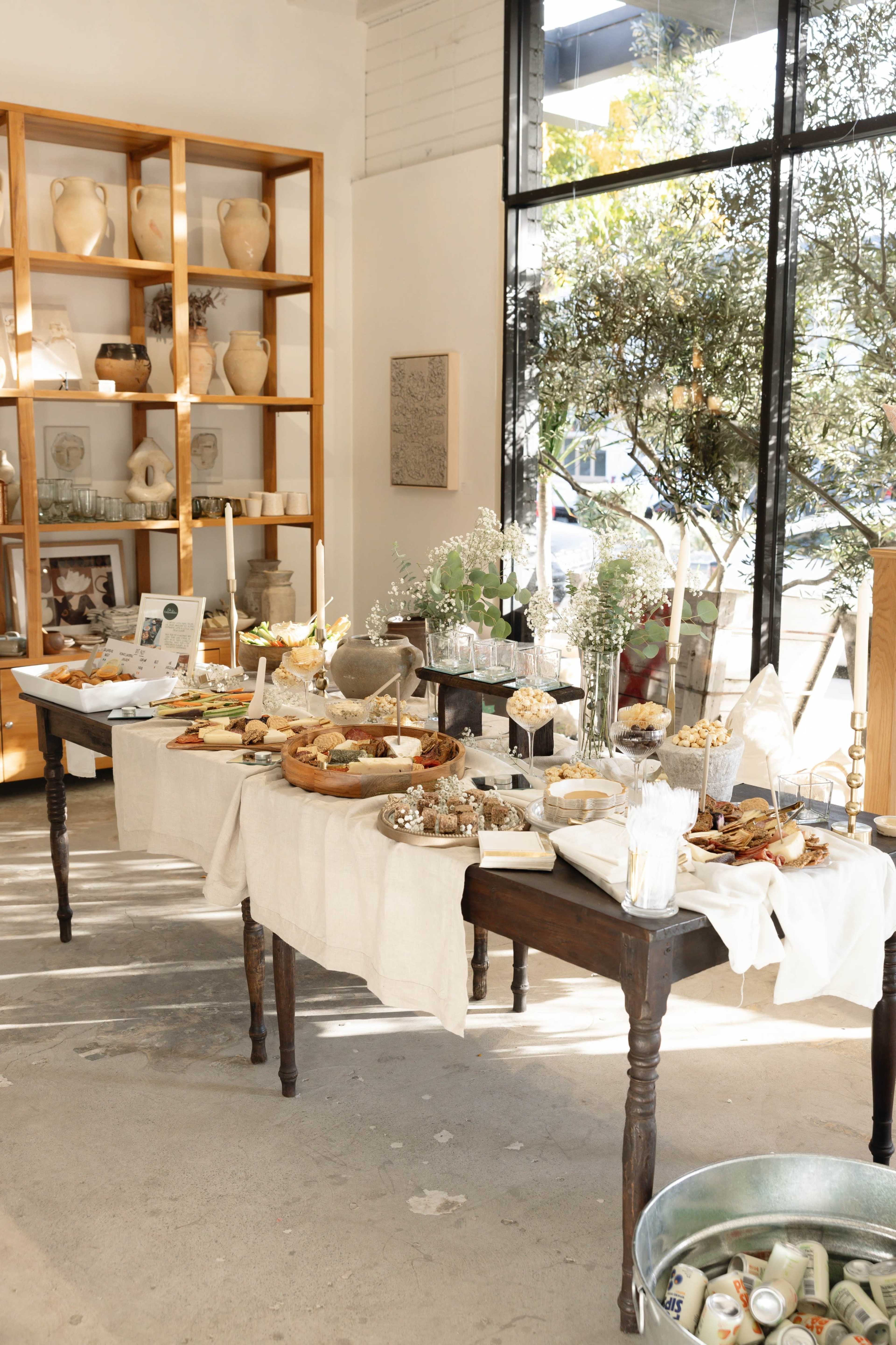 The scene features a beautifully arranged table with a variety of food items and decorative elements set in a well-lit room with large windows and a display of pottery in the background.