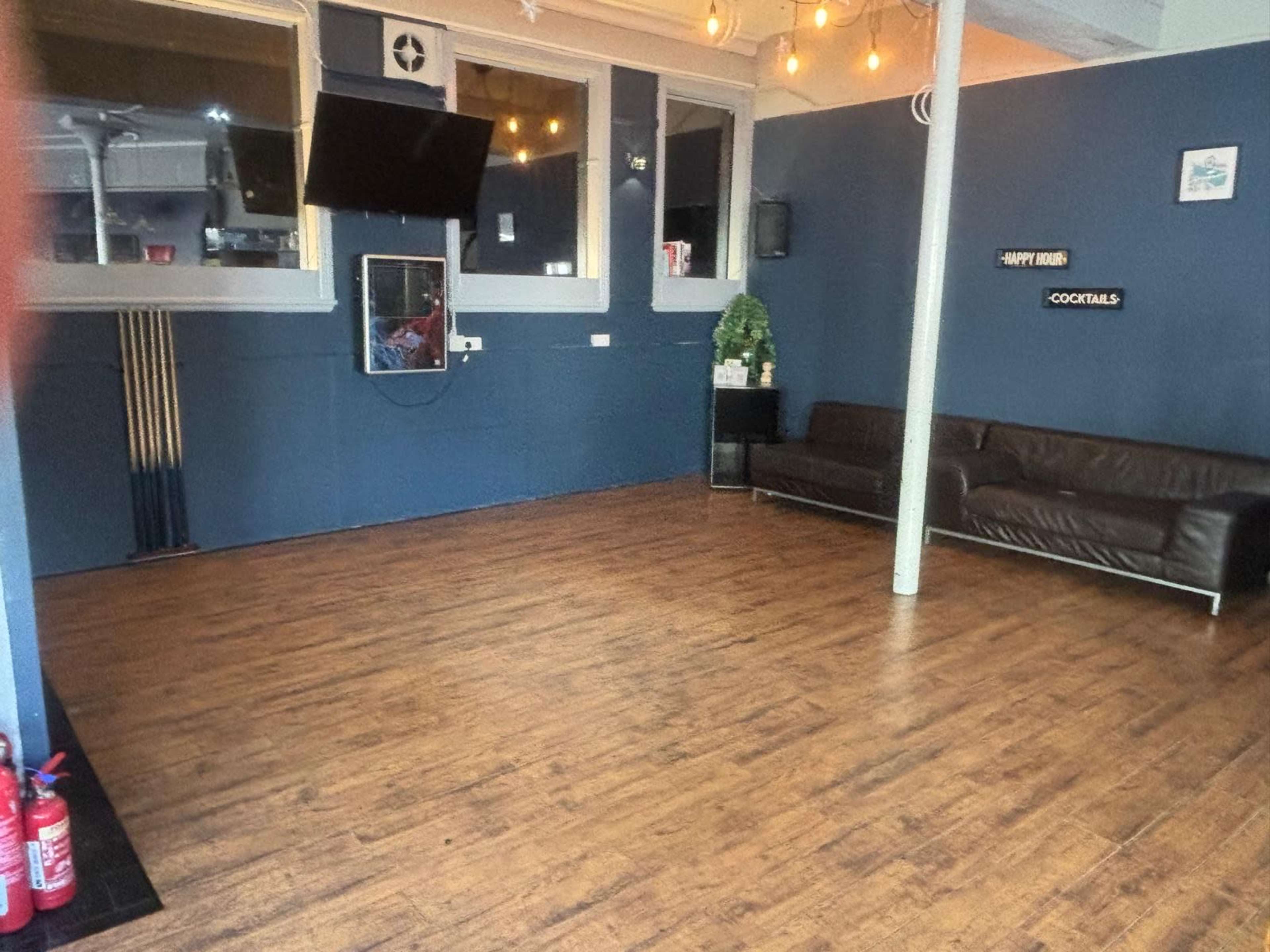 The image shows an empty room with a wooden floor, a dark leather sofa, and a wall featuring a sign that reads "COCKTAILS."