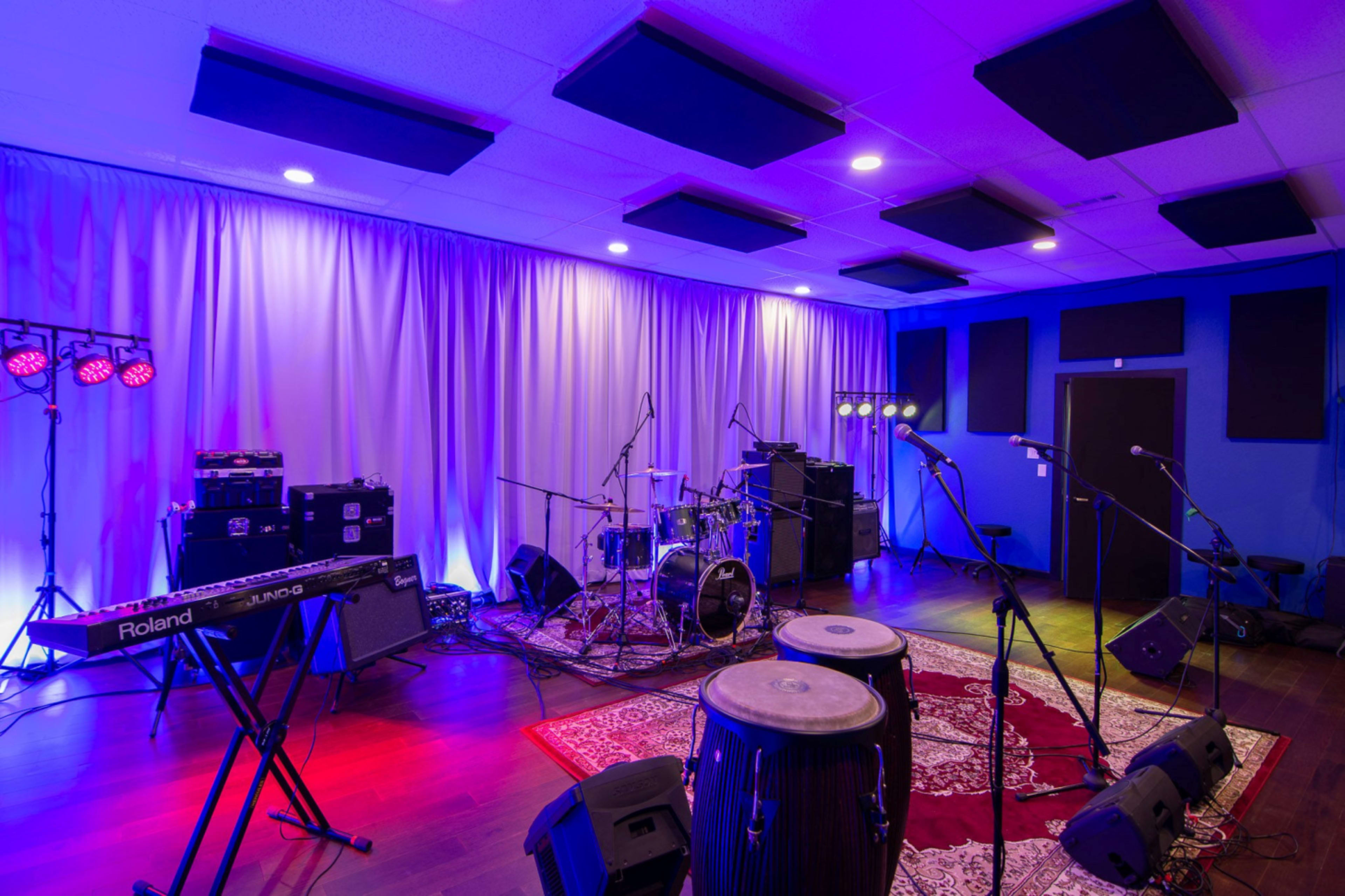 A music rehearsal studio is set up with various musical instruments, including microphones, a drum kit, keyboards, and amplifiers, illuminated by purple and blue lights.