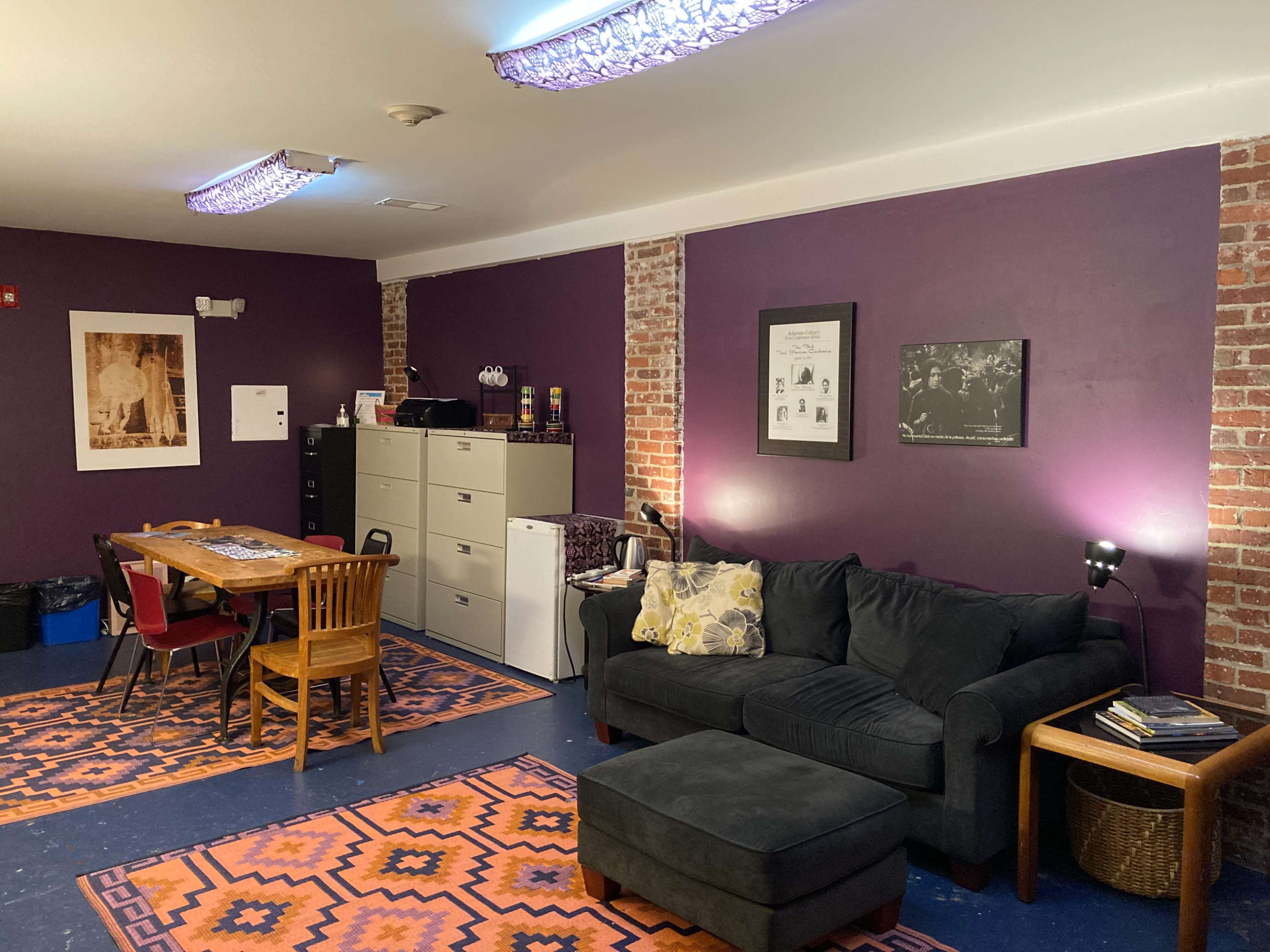 A room with purple walls, a dark sofa, a dining table with chairs, and filing cabinets, featuring patterned rugs on the floor.