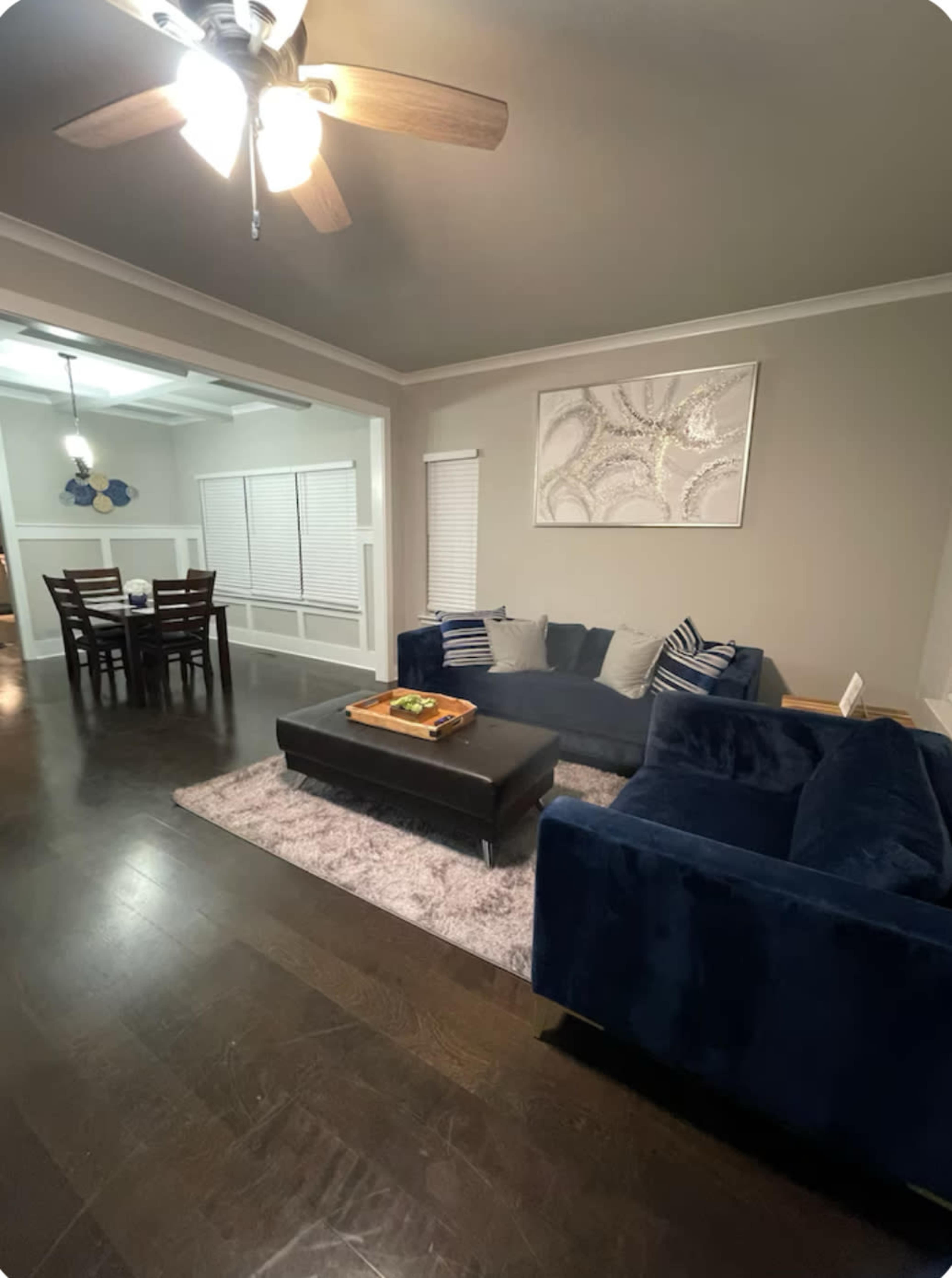A living room features a dark blue sectional sofa, a coffee table, and a light-colored rug, with a dining area visible in the background.