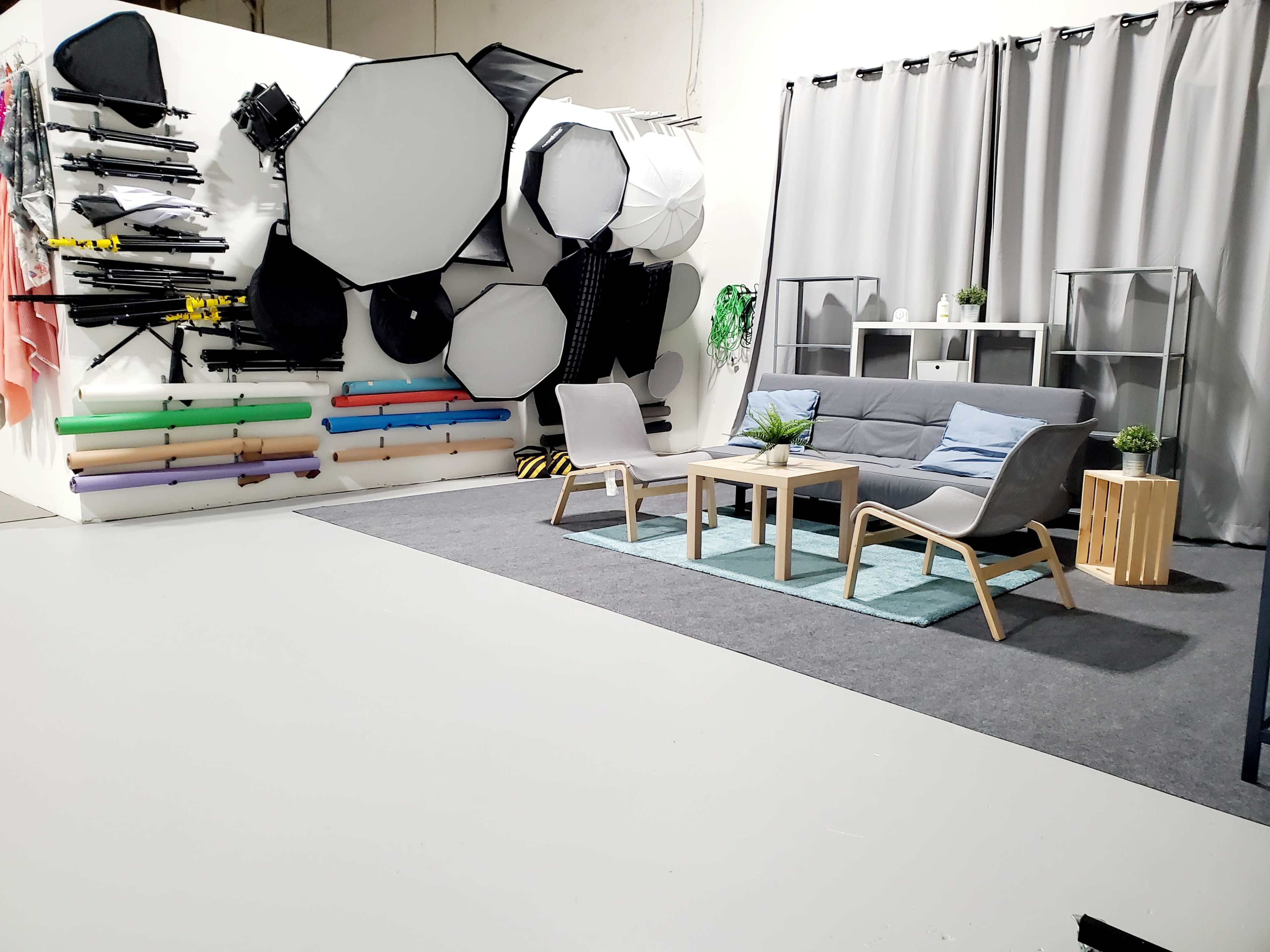 A photography studio setup featuring various backdrops and lighting equipment on the wall, a gray sofa, a coffee table, and a rug in a minimalistic layout.