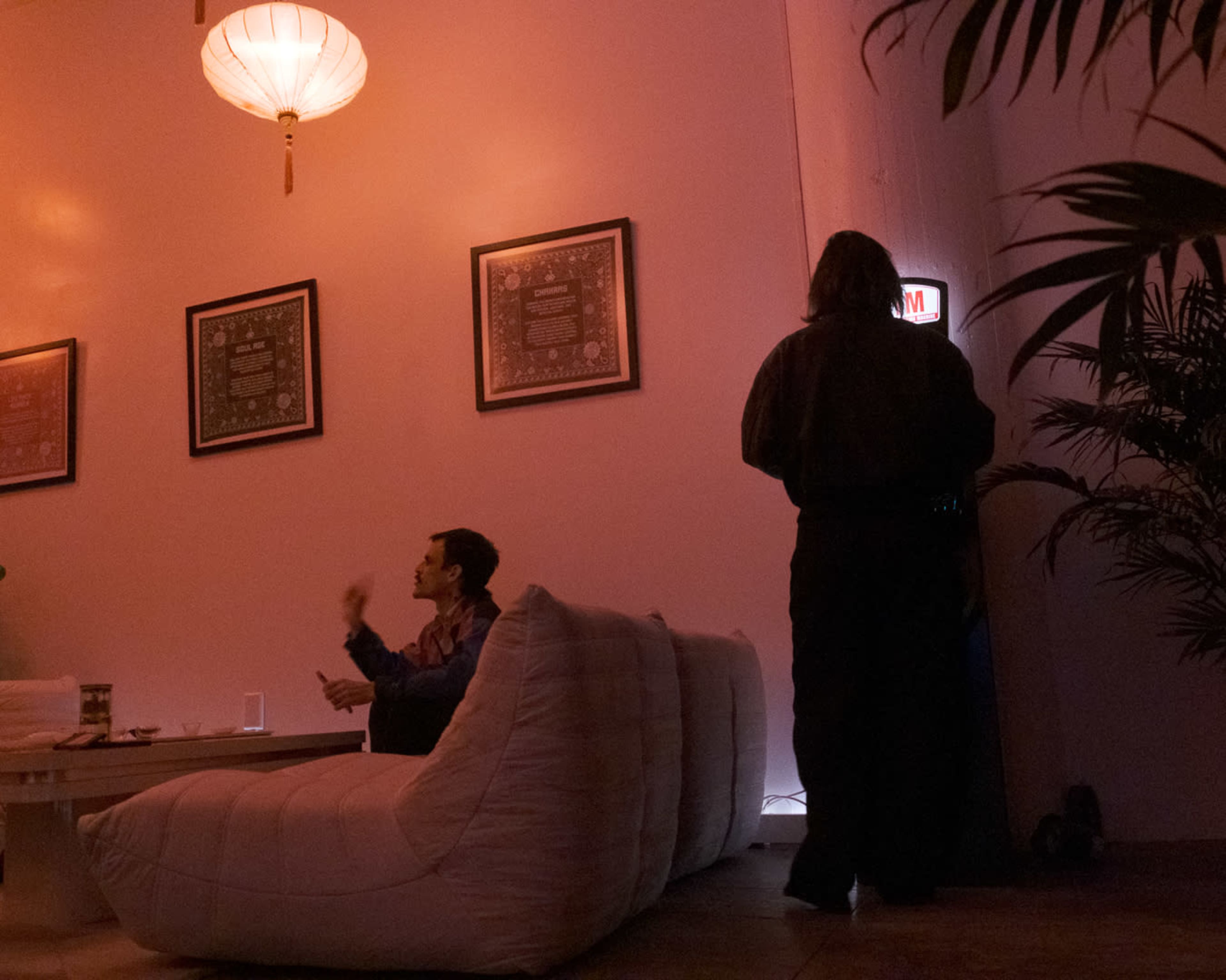 A dimly lit room features two individuals, one sitting on a large white couch and the other standing near a door illuminated by a red "Open" sign.