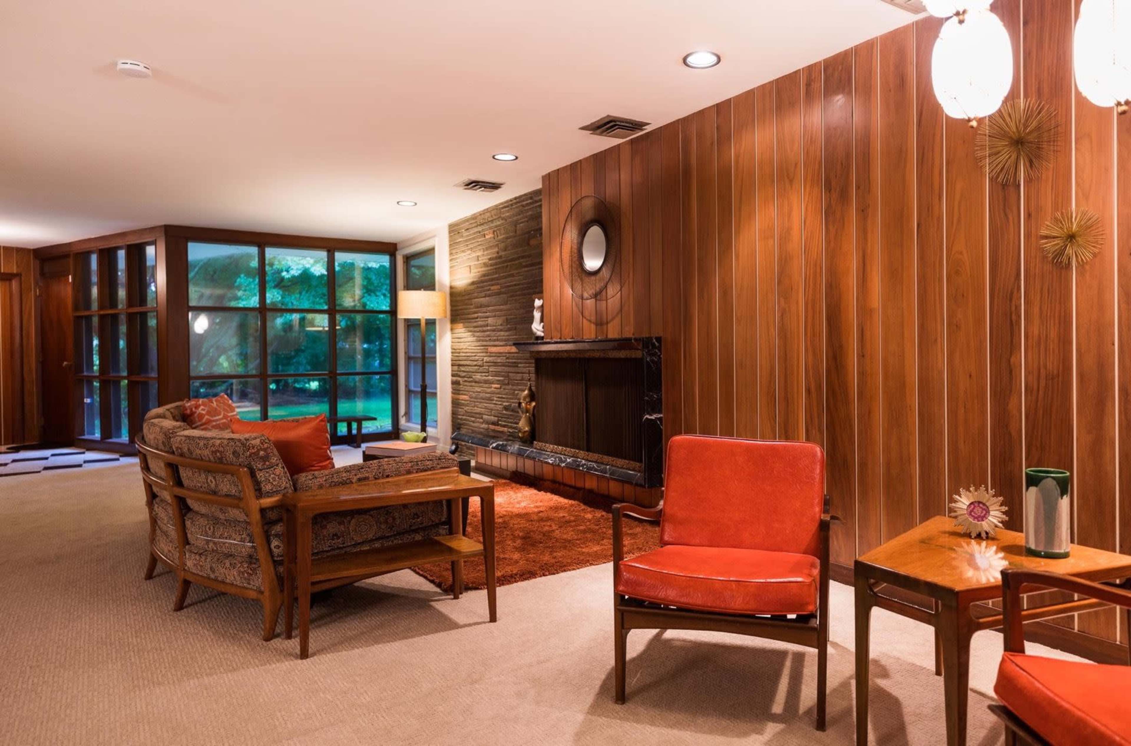 The image displays a mid-century modern living room featuring wooden paneling, a fireplace, and vintage furniture arrangements on carpeted flooring.