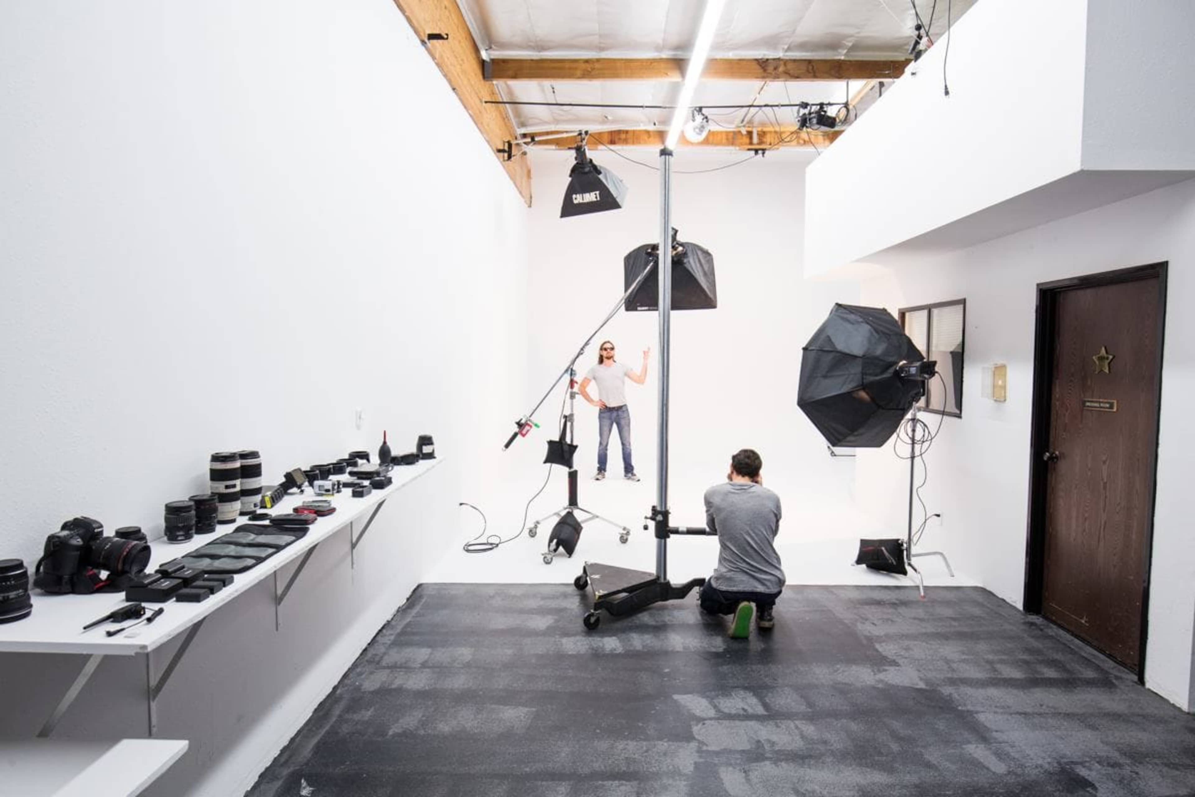 A photographer is capturing a model's portrait in a well-lit studio, equipped with various cameras and lenses displayed on a shelf to the left.