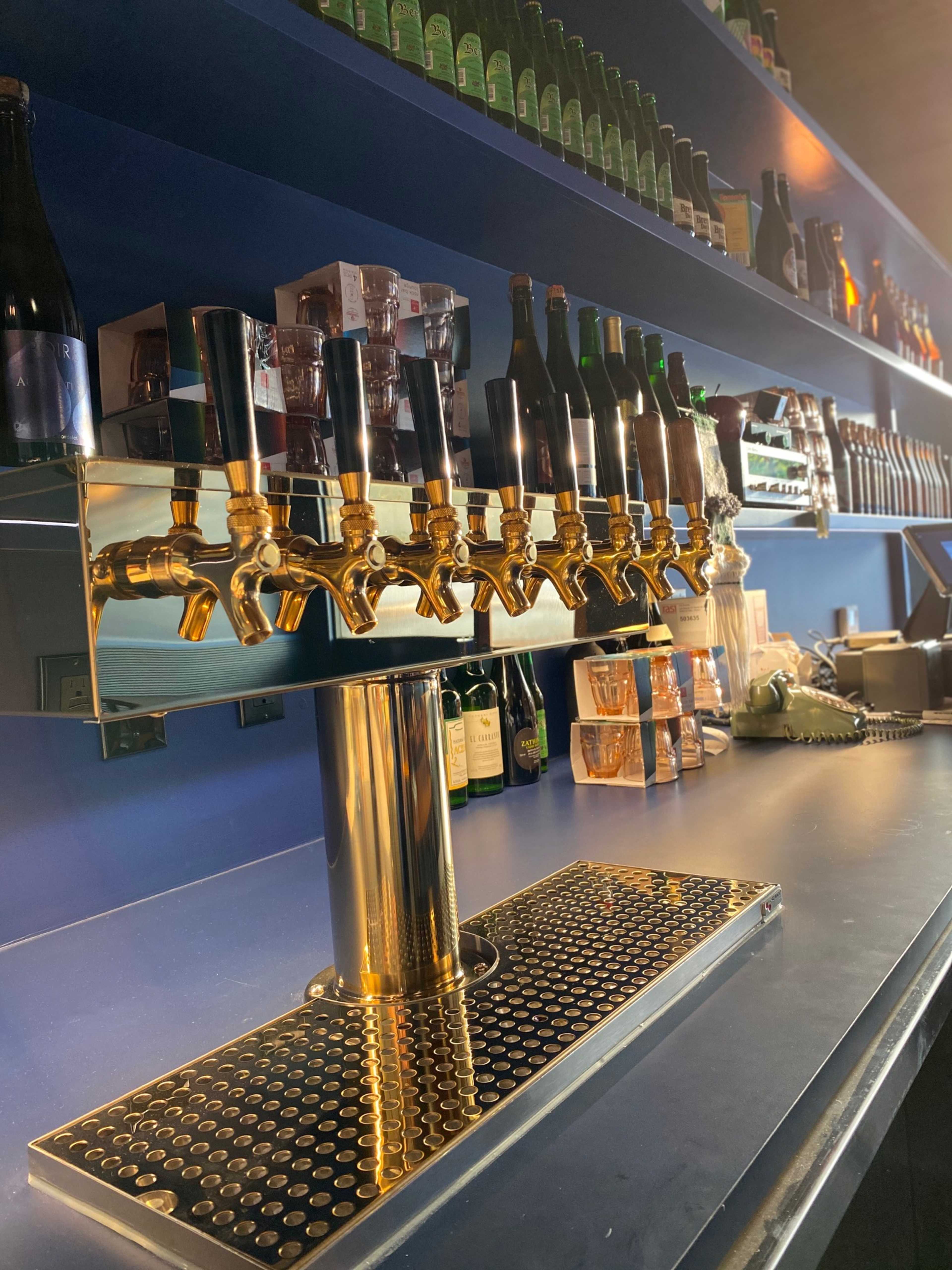 A polished bar with a row of eight shiny brass beer taps and shelves lined with various bottles in the background.