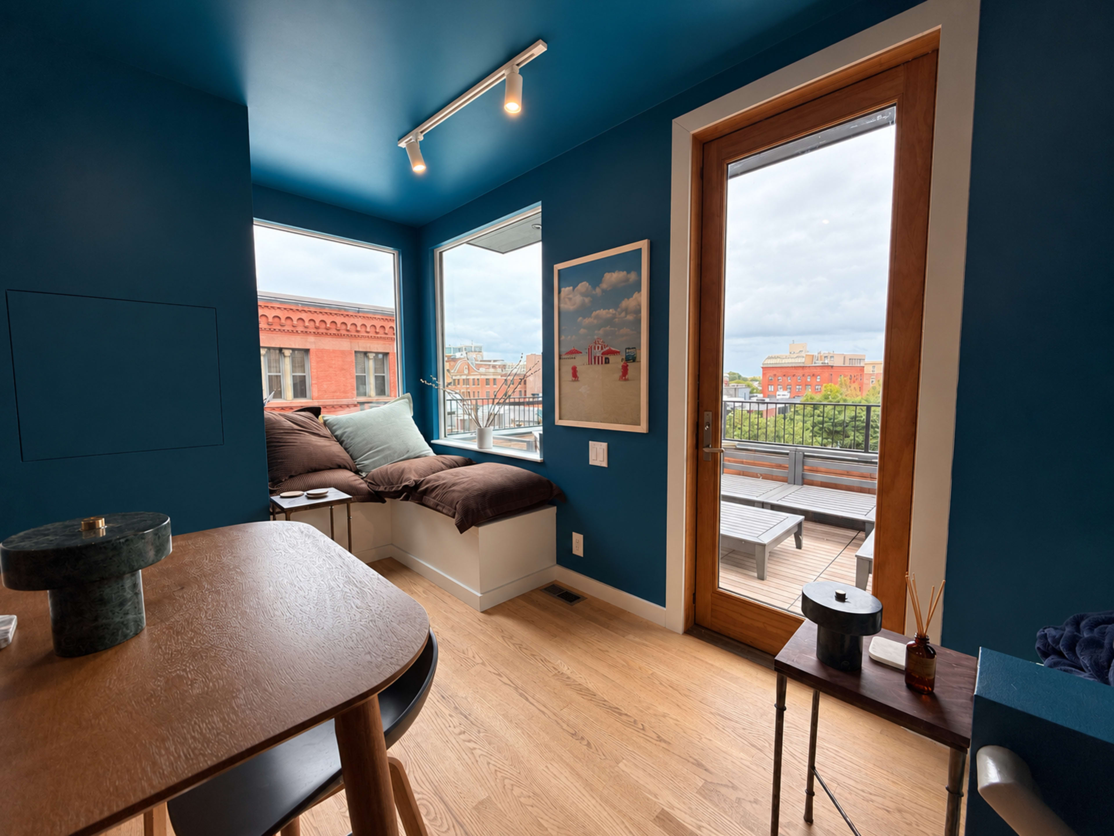 A cozy room features a blue wall, a window seat with cushions, and a door leading to a rooftop terrace.