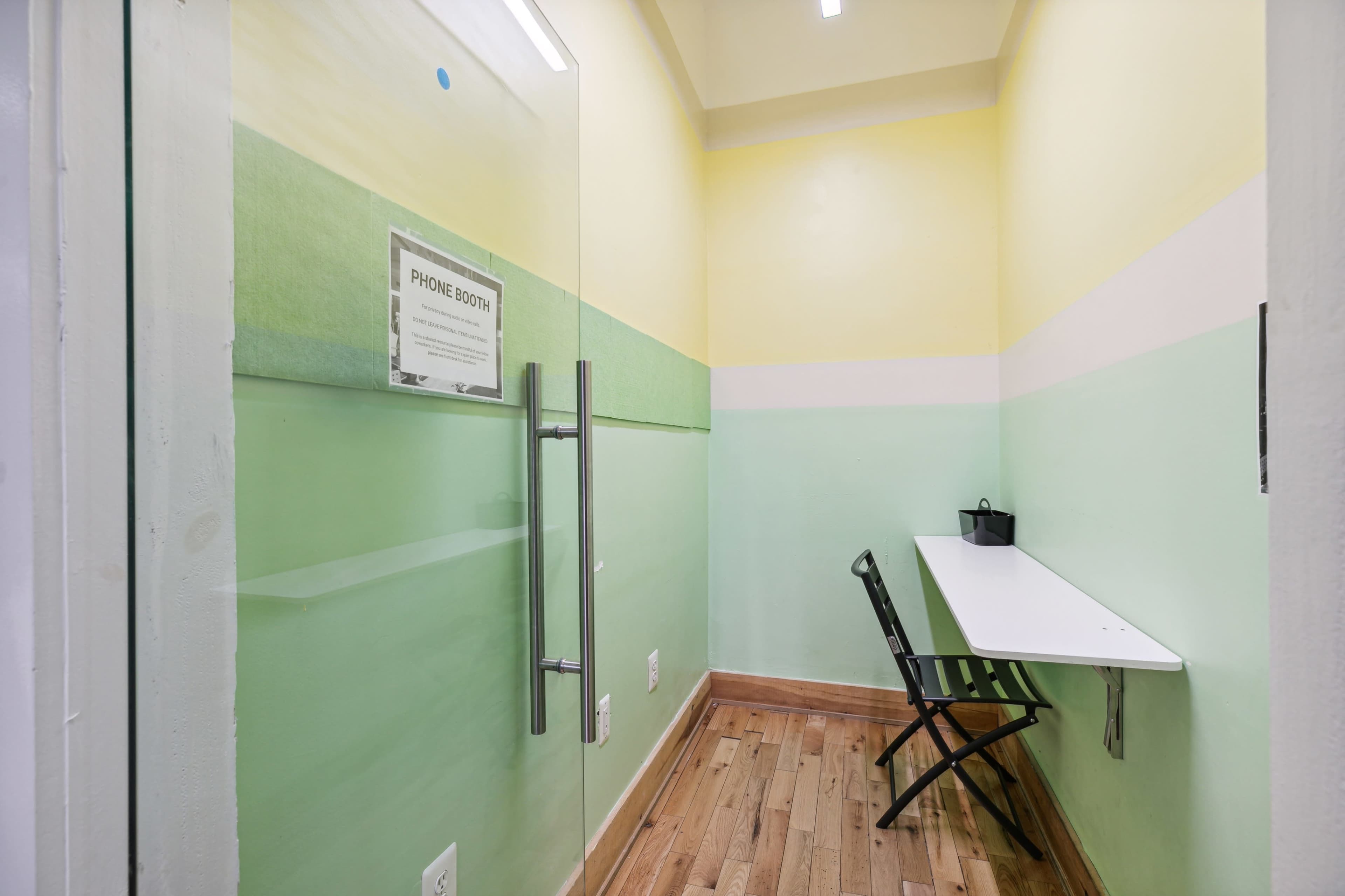 A small phone booth with a glass door, a white wall-mounted desk, and a folding chair.
