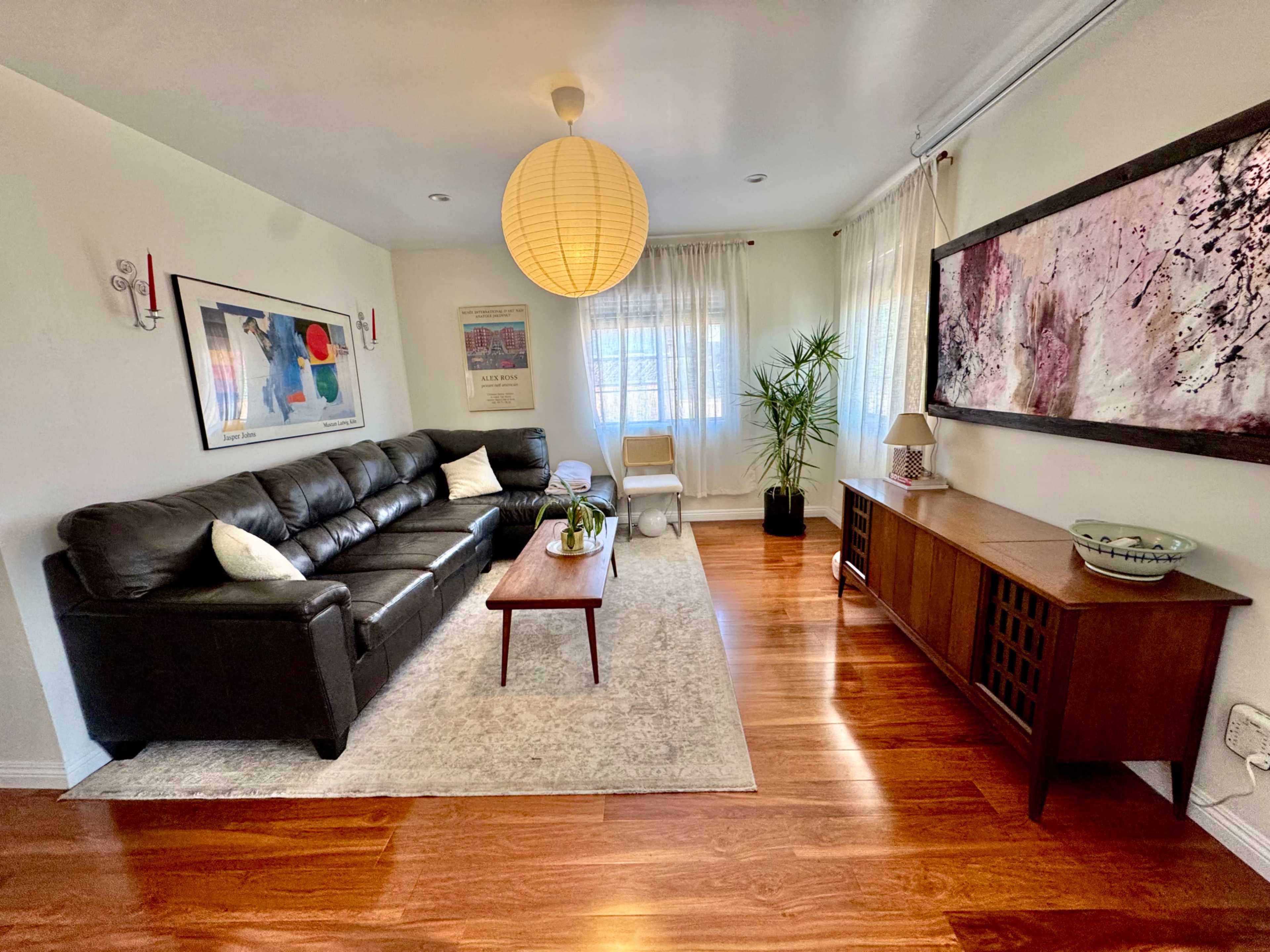The image shows a modern living room featuring a black leather sofa, wooden flooring, a coffee table, and a decorative wall painting.