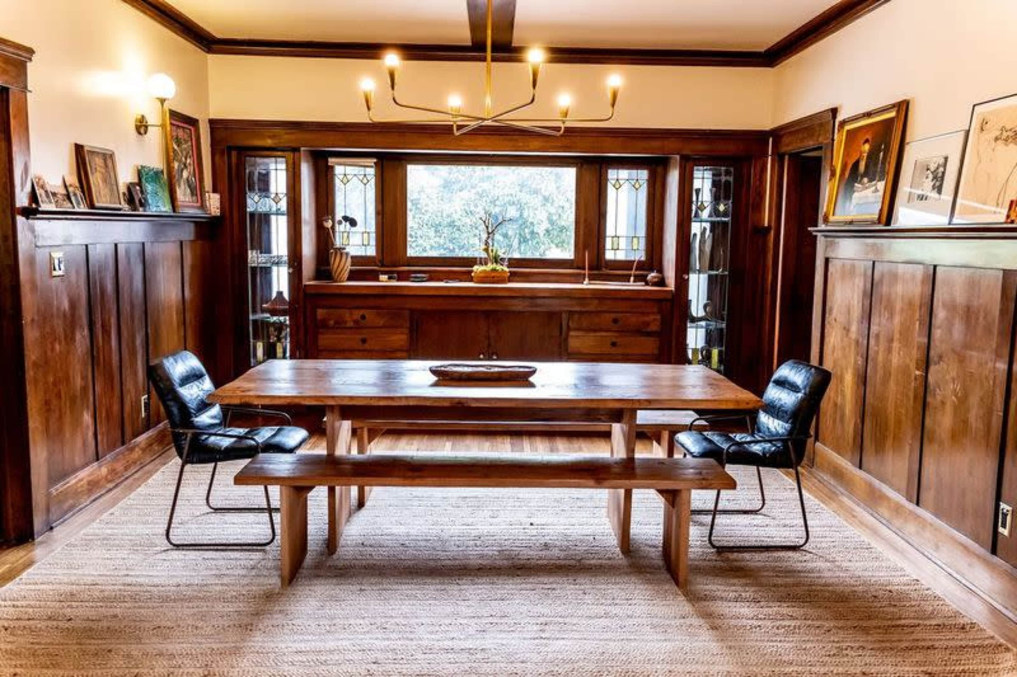 A wooden dining table with a bench and two black chairs is positioned in a room with dark paneling and large windows.