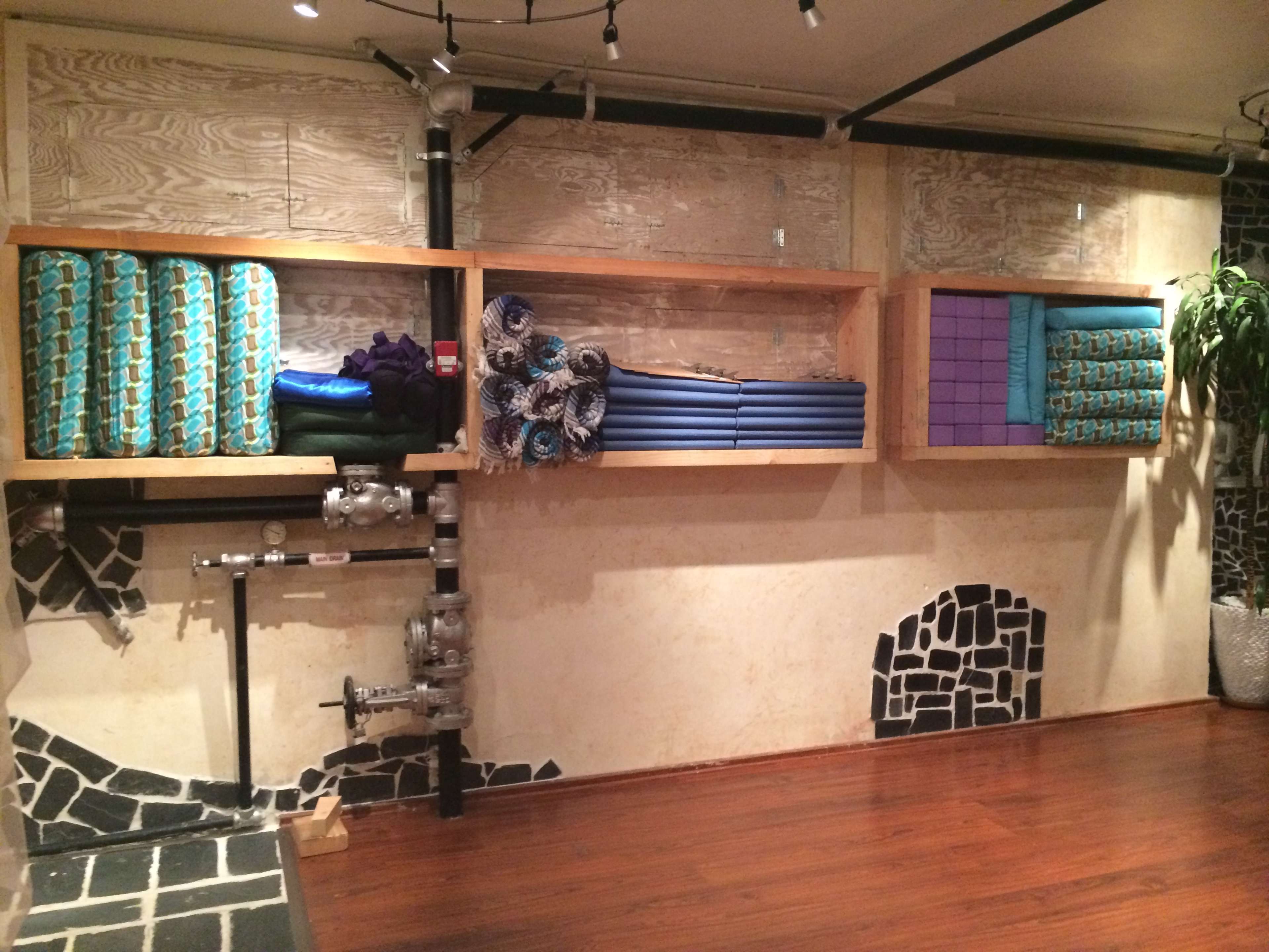 The image shows a wall with wooden shelving holding neatly rolled yoga mats and exercise equipment, alongside a pipe installation and a potted plant.