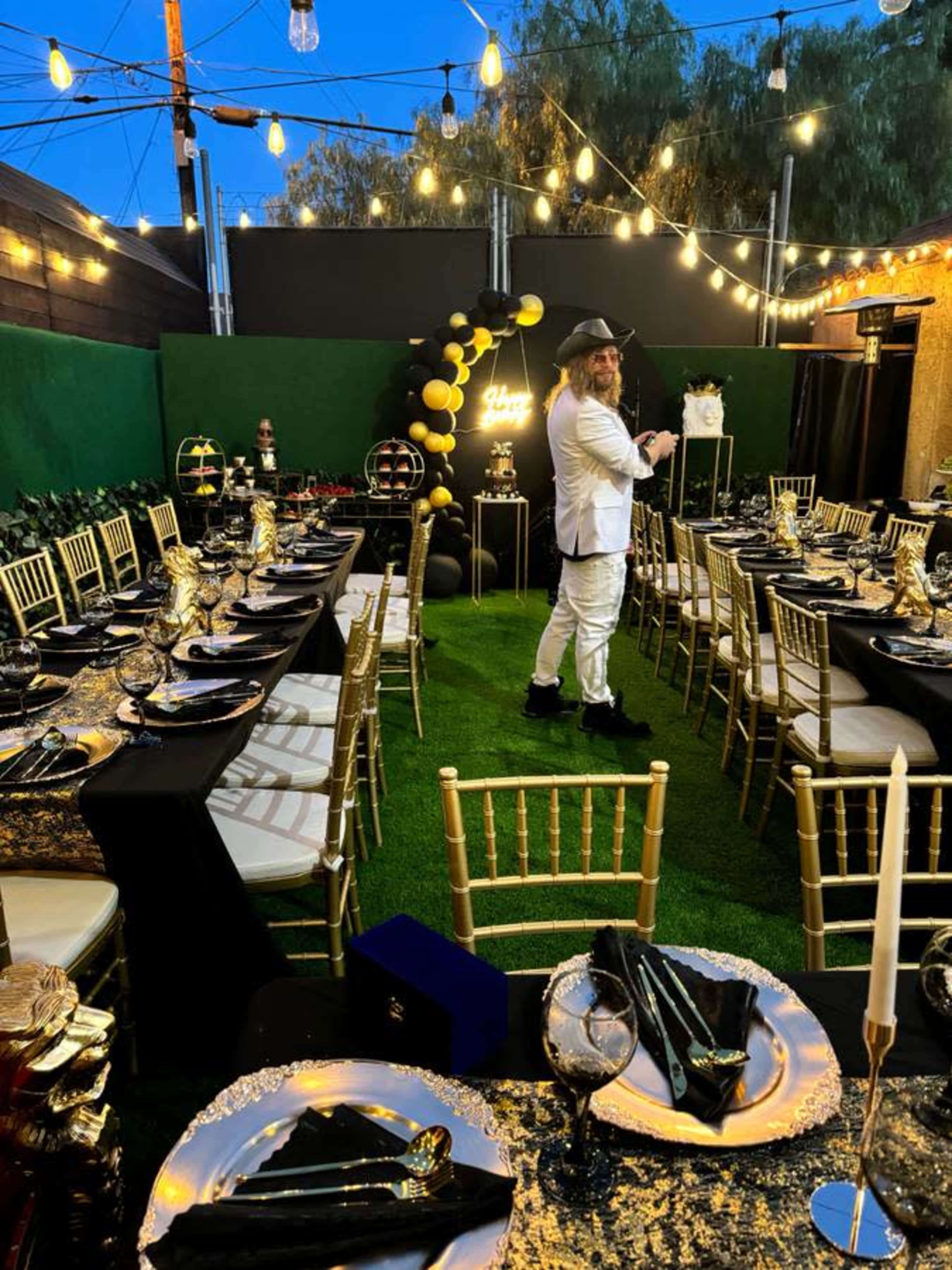 A man in a white suit stands in a decorated outdoor dining area with gold and black party decor, surrounded by tables set with elegant tableware and string lights overhead.
