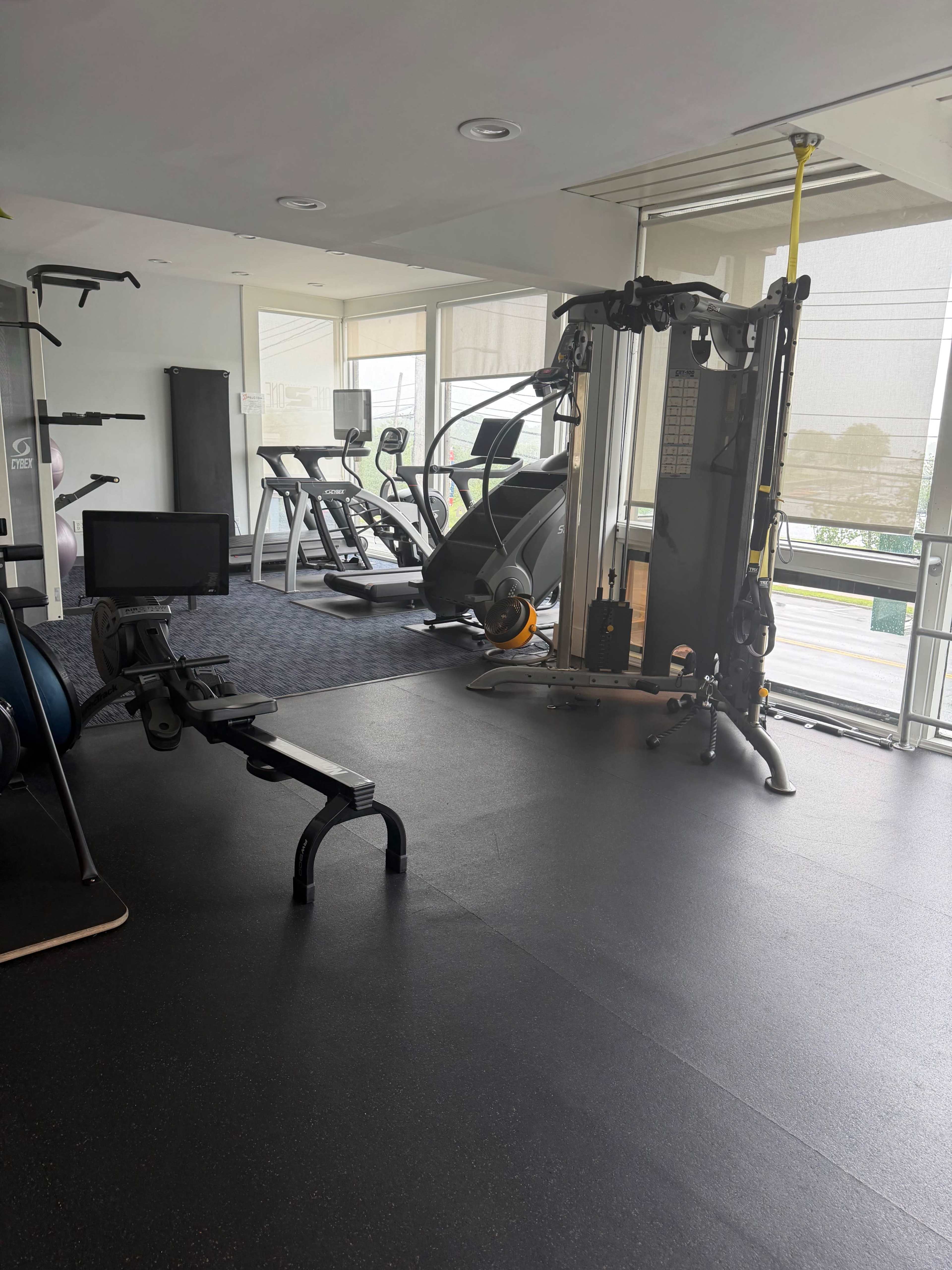 The image shows a modern gym interior featuring various exercise equipment, including a cable machine, treadmill, elliptical, and weight bench.