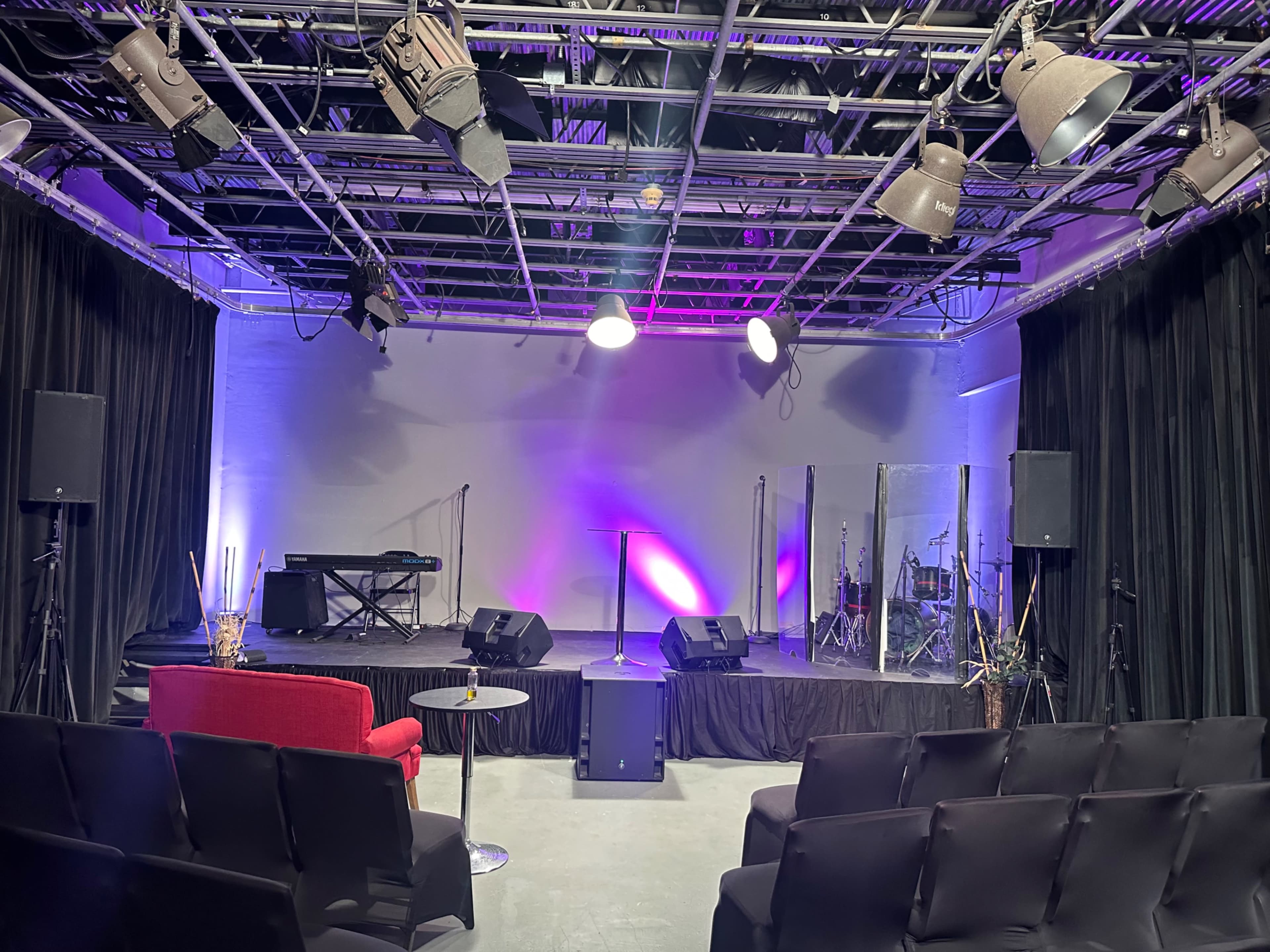 The image shows a performance space with a stage, musical instruments, and seating arranged in front of a black backdrop.