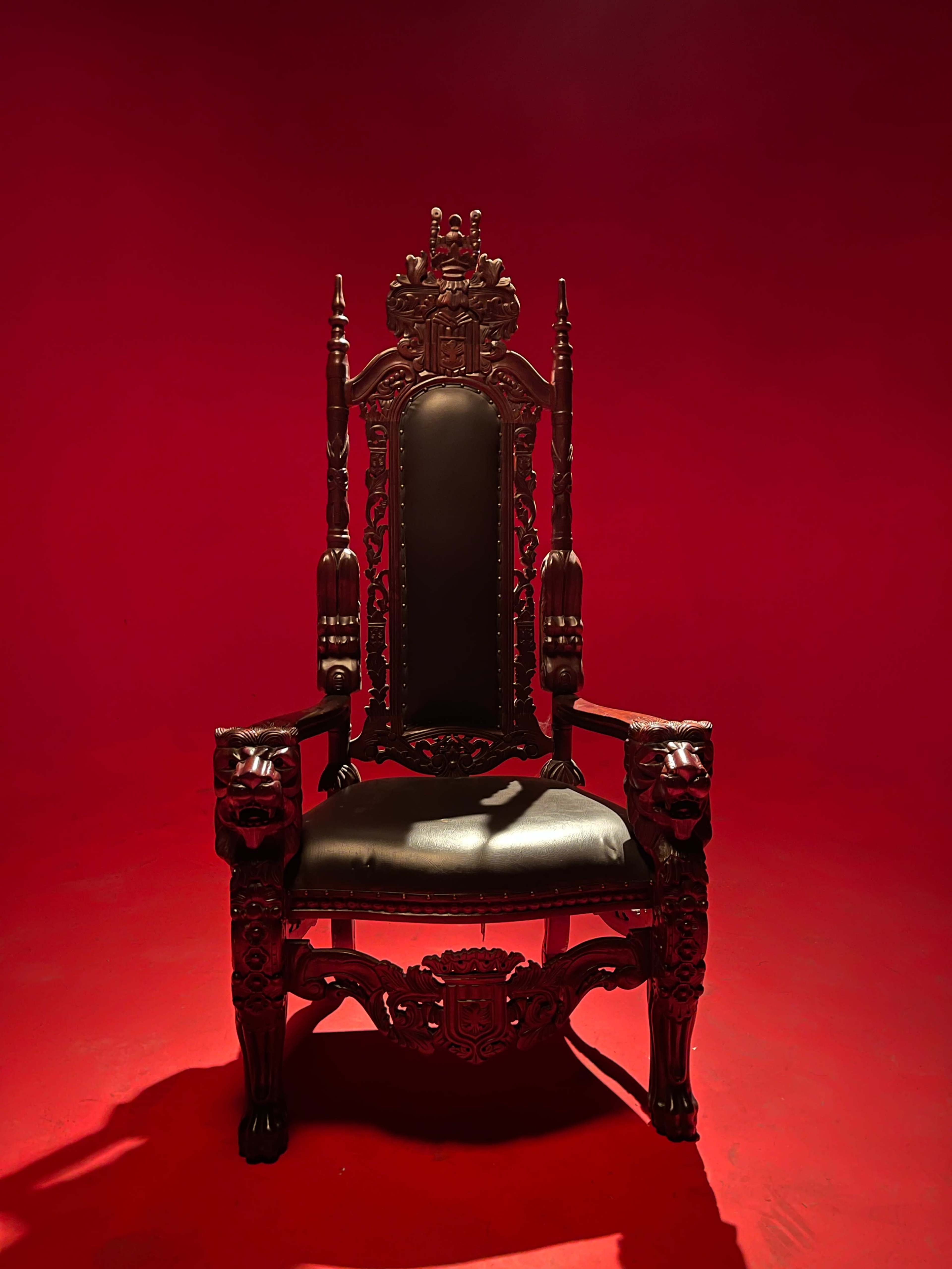 A tall, ornate wooden throne with lion head armrests is positioned in front of a solid red backdrop.
