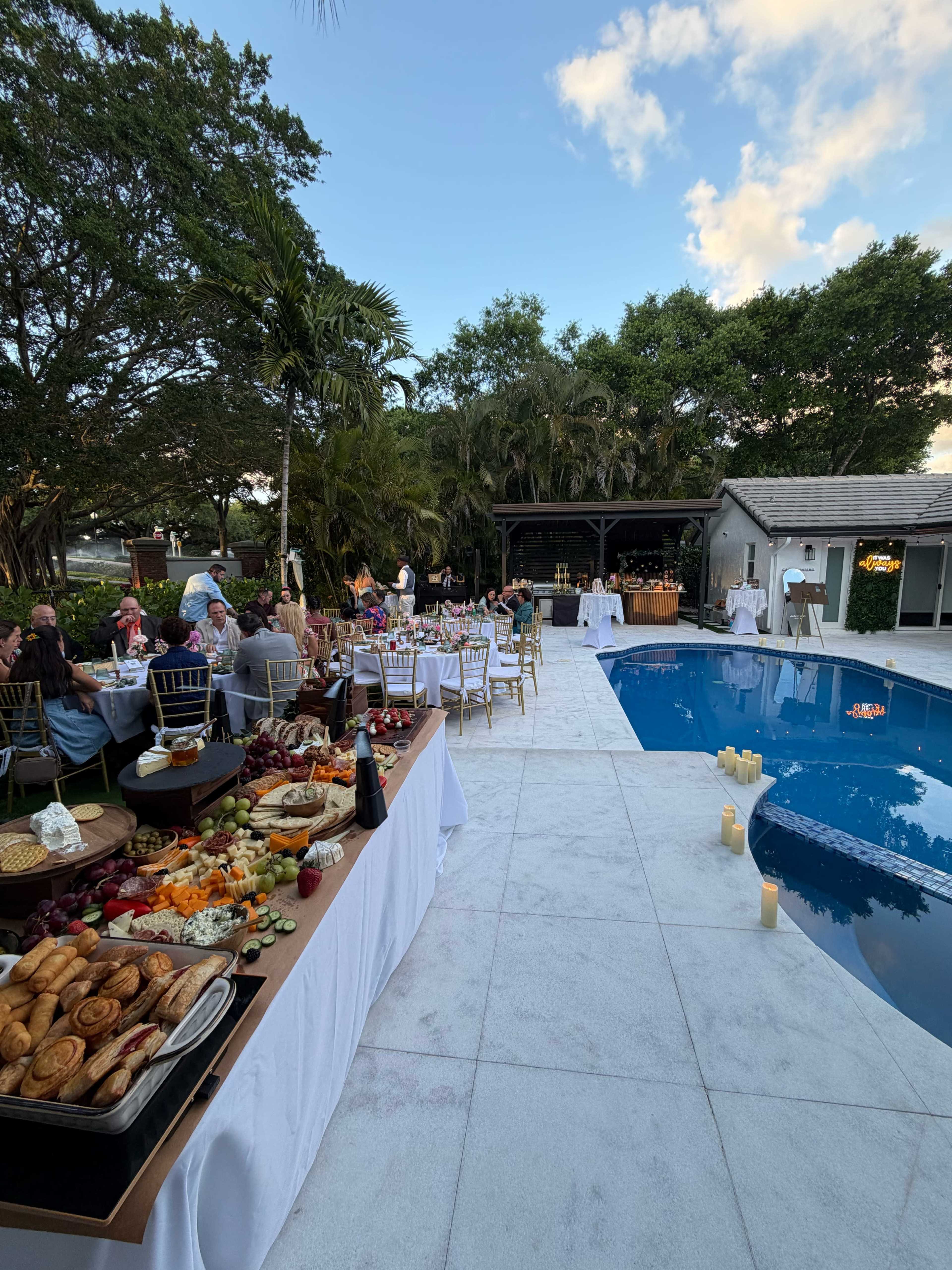 A poolside gathering features elegantly arranged tables with food and drinks, surrounded by greenery and guests enjoying the outdoor atmosphere.