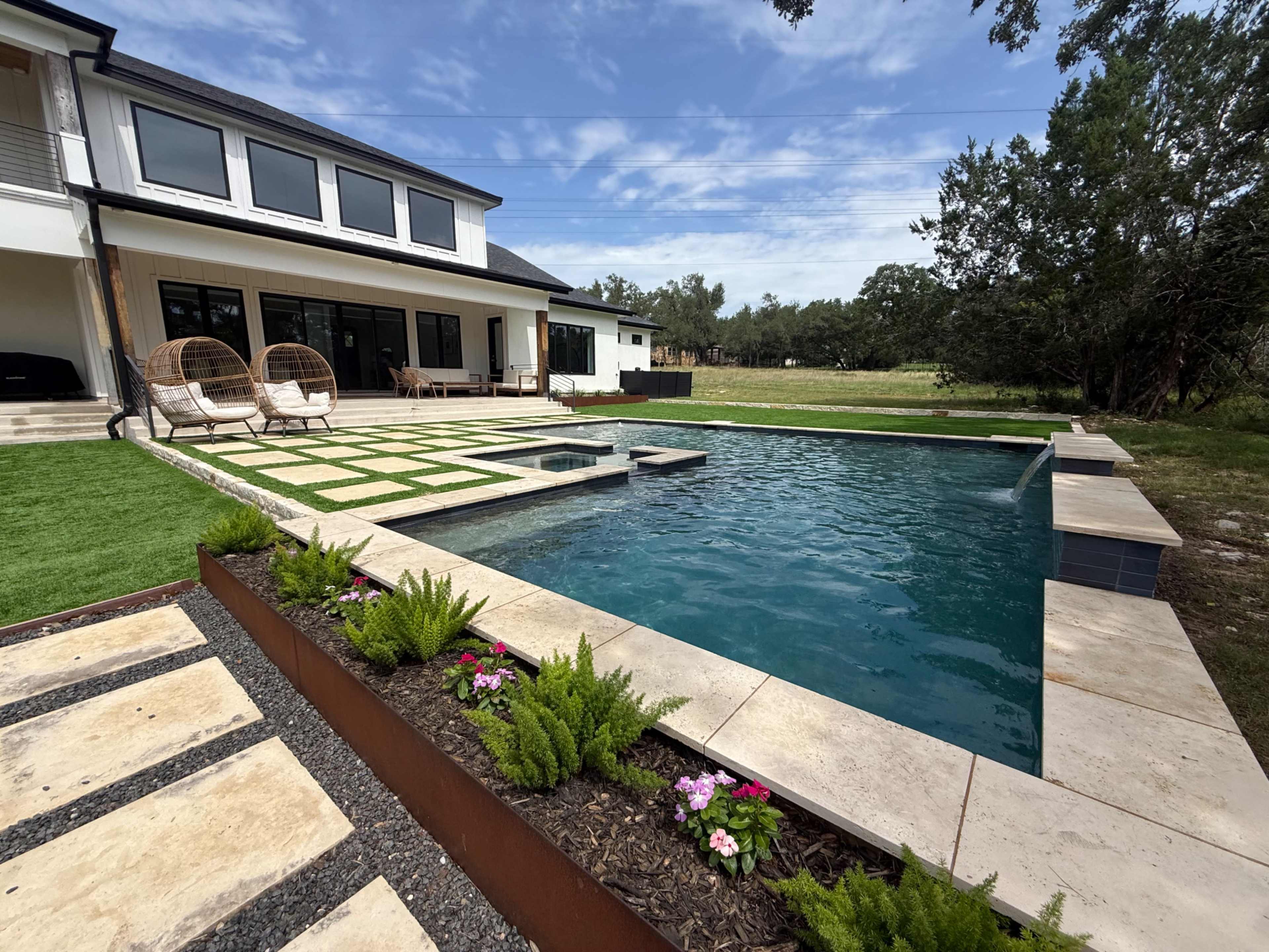 The image shows a modern backyard featuring a rectangular swimming pool surrounded by landscaped areas, including green grass and flower beds, with lounge chairs nearby.