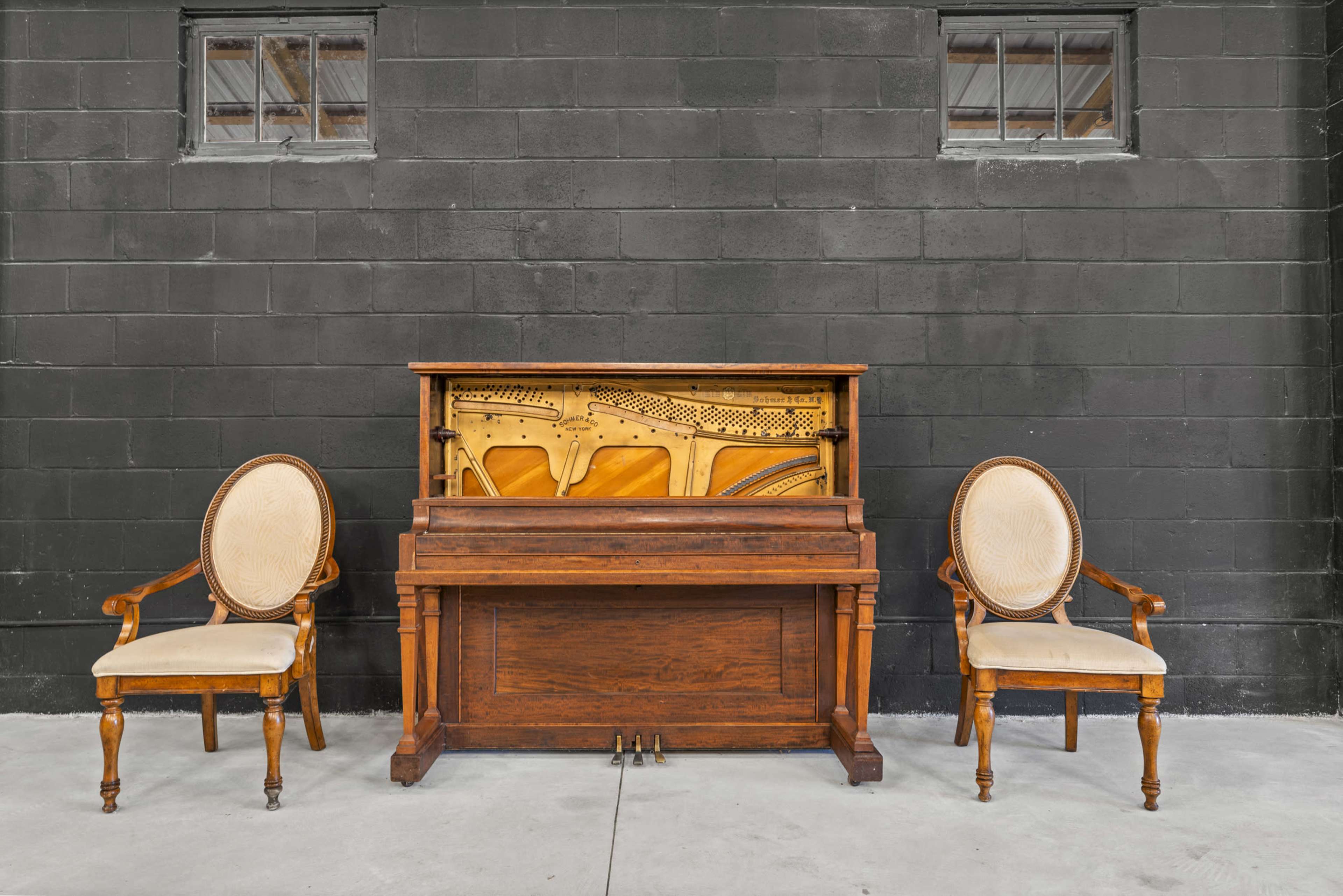 An upright piano is placed between two vintage chairs against a black brick wall.