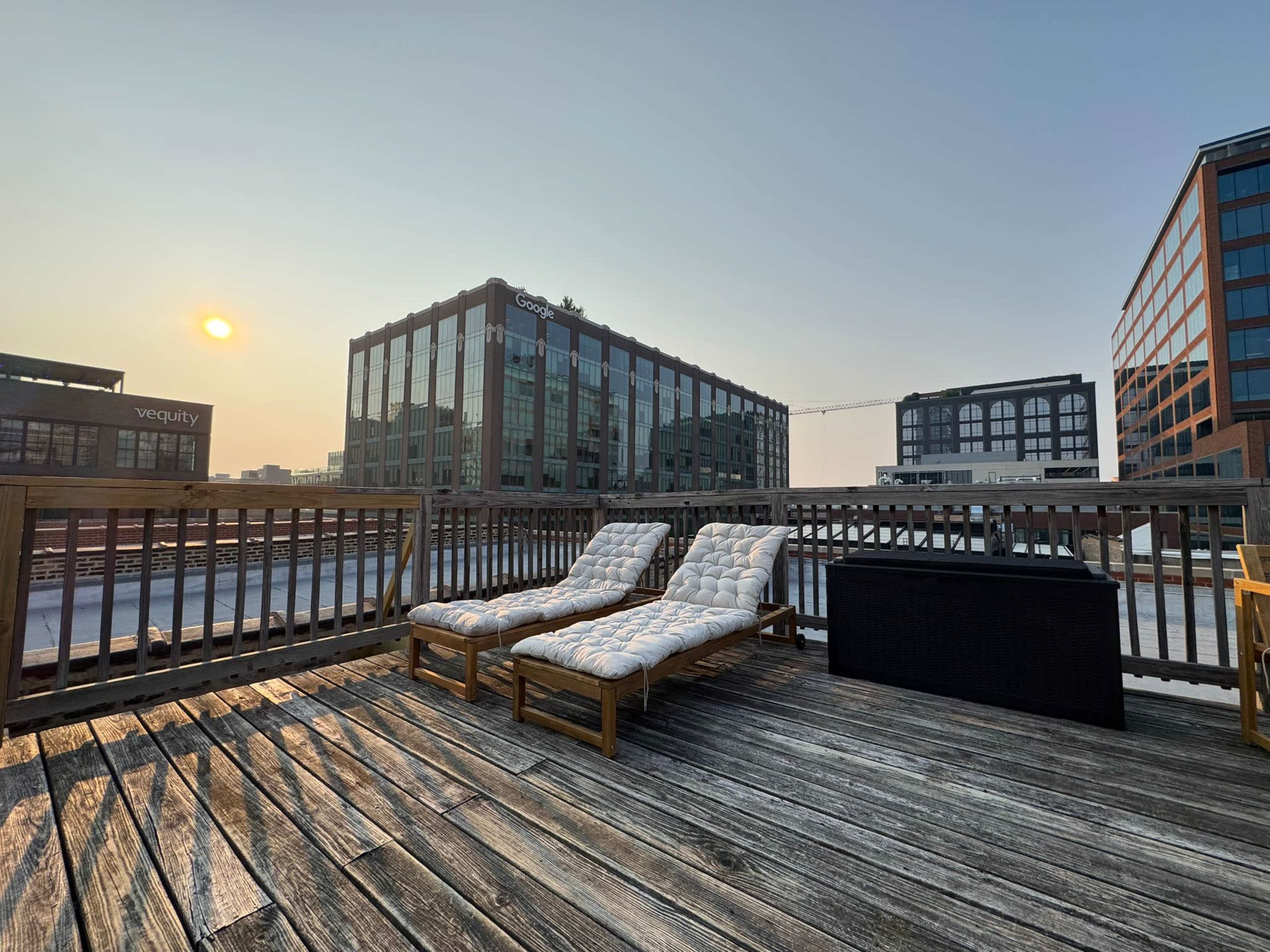 The image shows two lounge chairs on a wooden deck with modern buildings and a setting sun in the background.