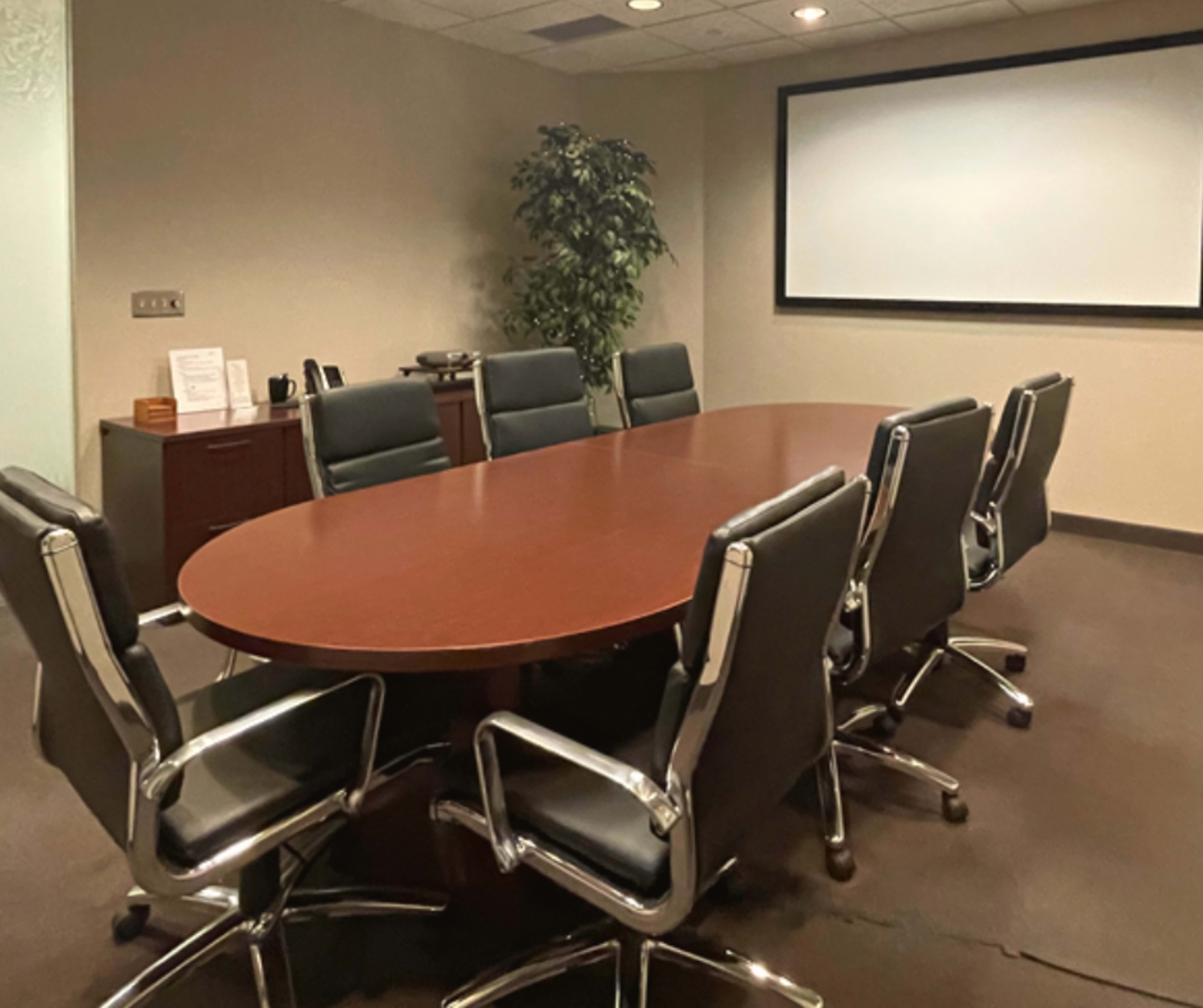 A conference room features a large oval wooden table surrounded by eight black leather chairs and a screen on the wall.