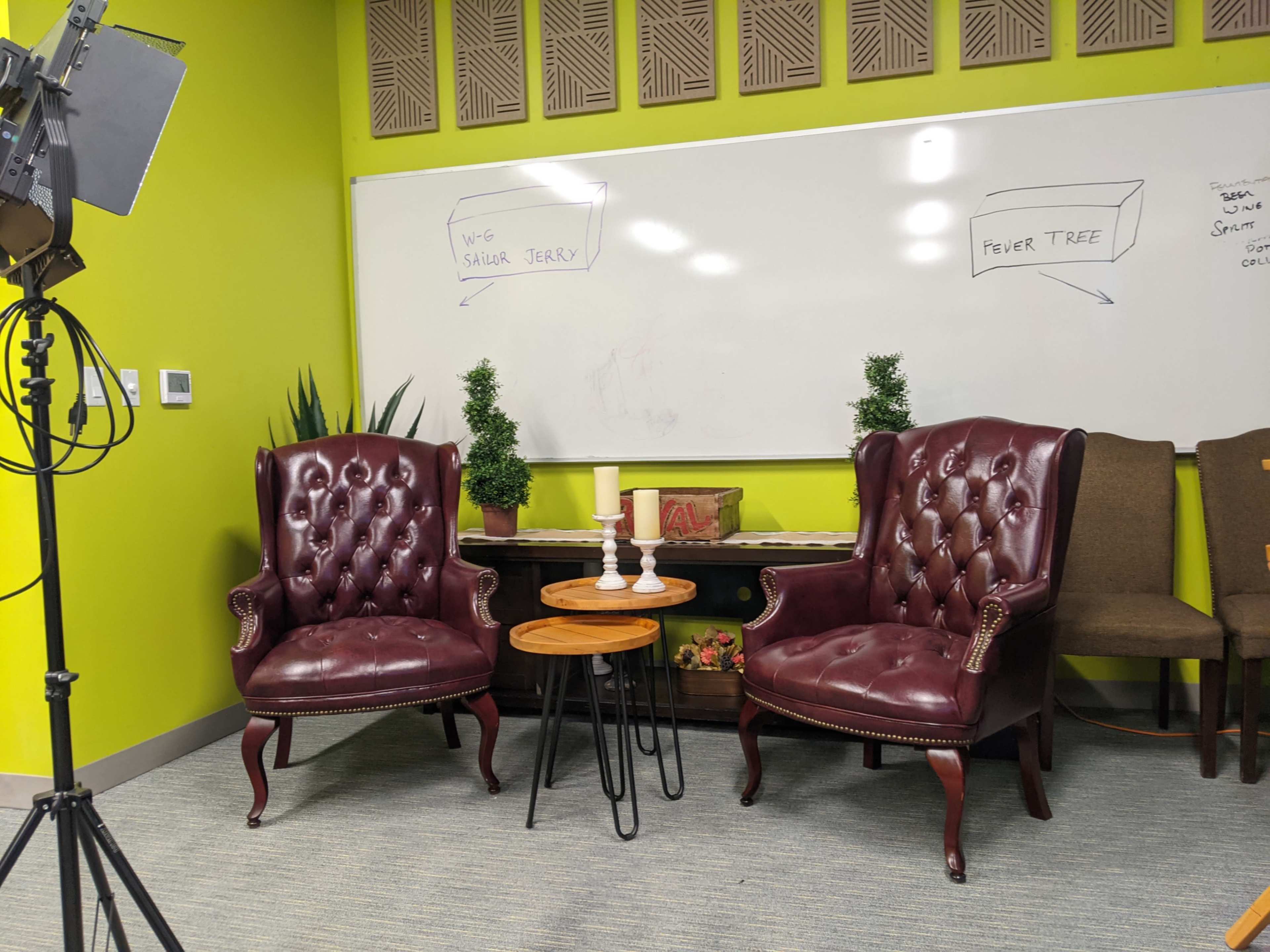 The image shows a small interview or discussion setup with two burgundy upholstered chairs facing a wooden table, positioned in front of a whiteboard against a bright green wall.