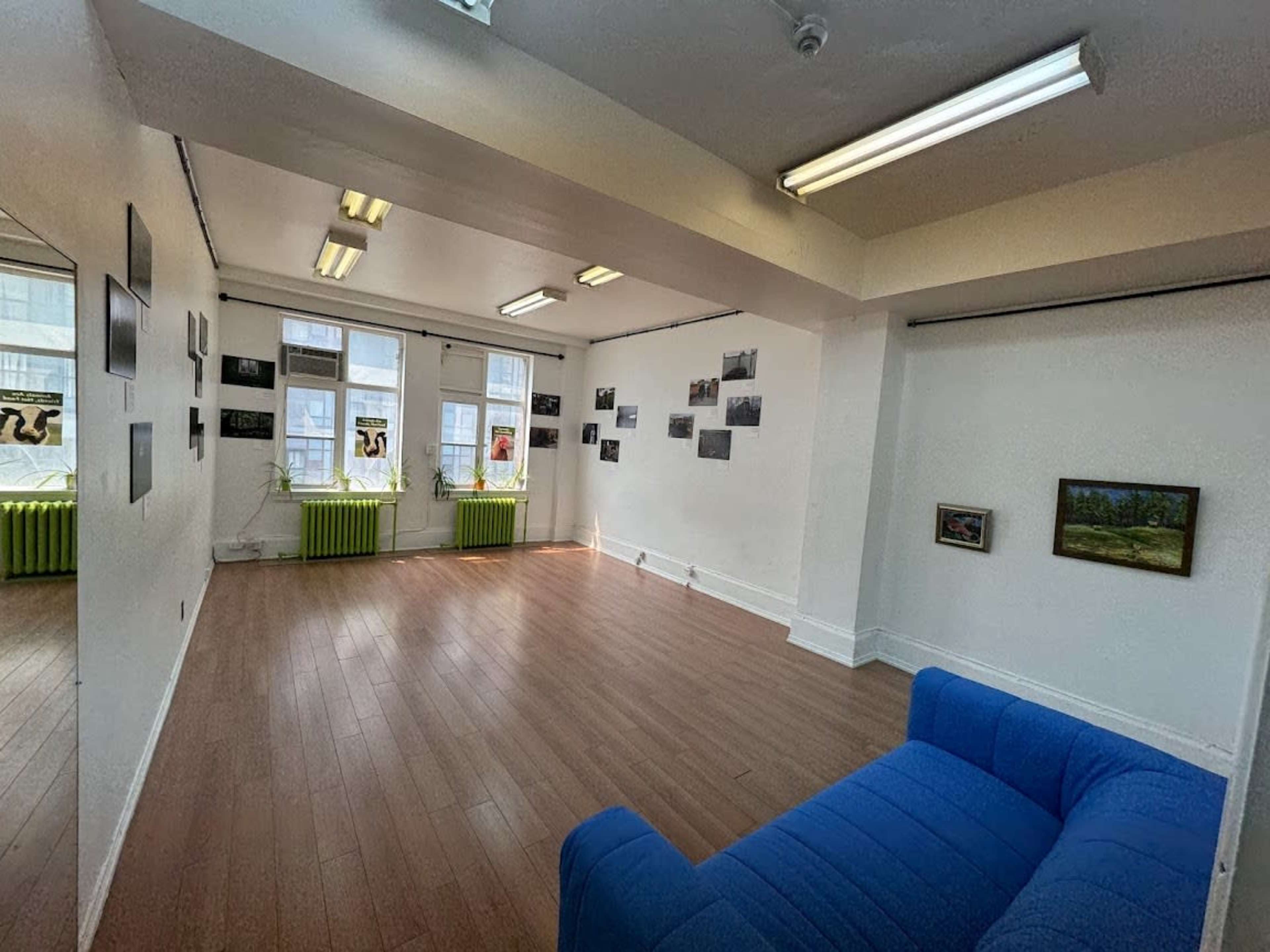 The image shows a spacious, well-lit interior of an art gallery or studio with wooden flooring, featuring a blue couch and several framed pictures on the walls.