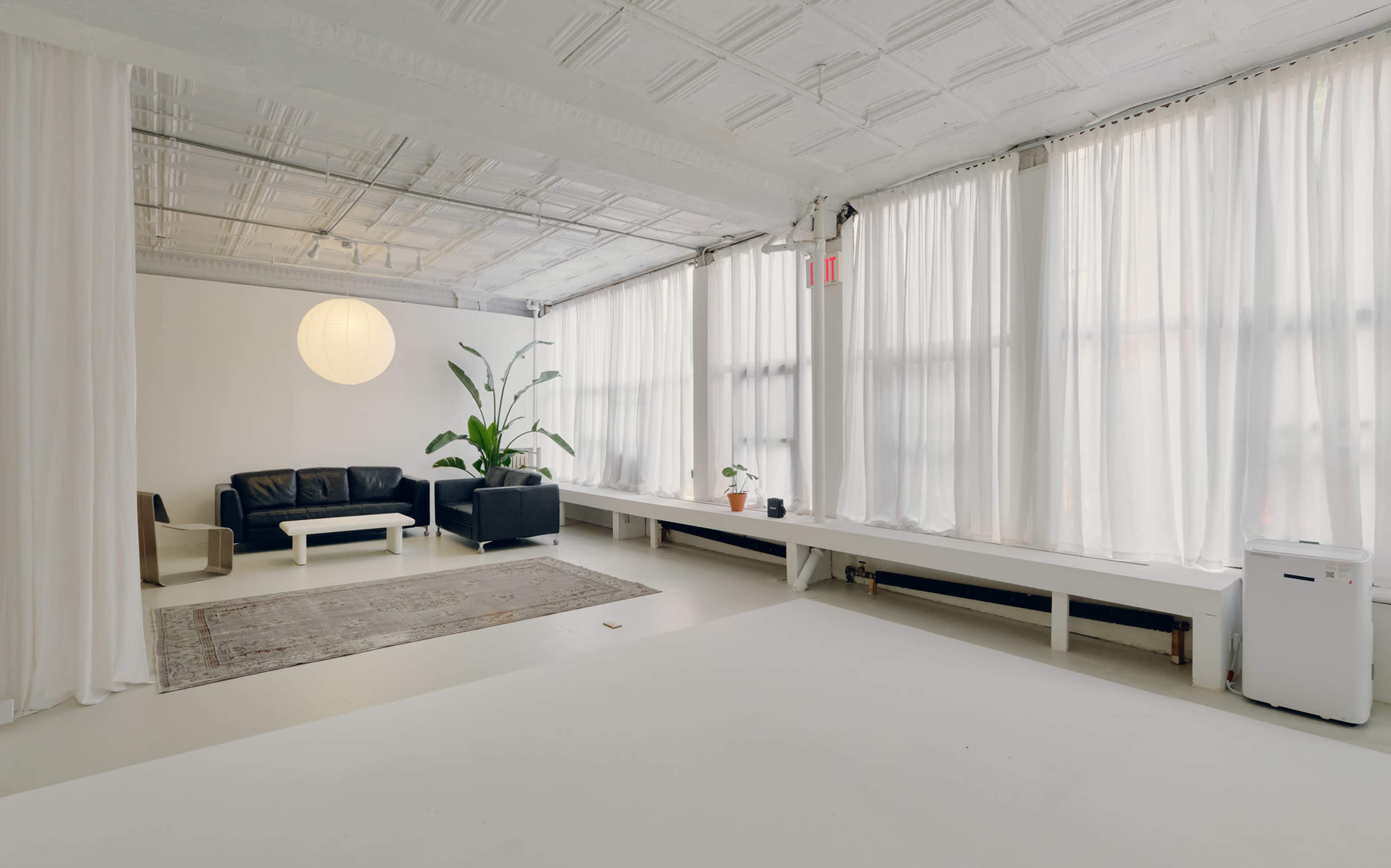 The image shows a spacious, minimalistic room with large windows, a black sofa, a chair, and potted plants, along with a textured ceiling and light-colored flooring.