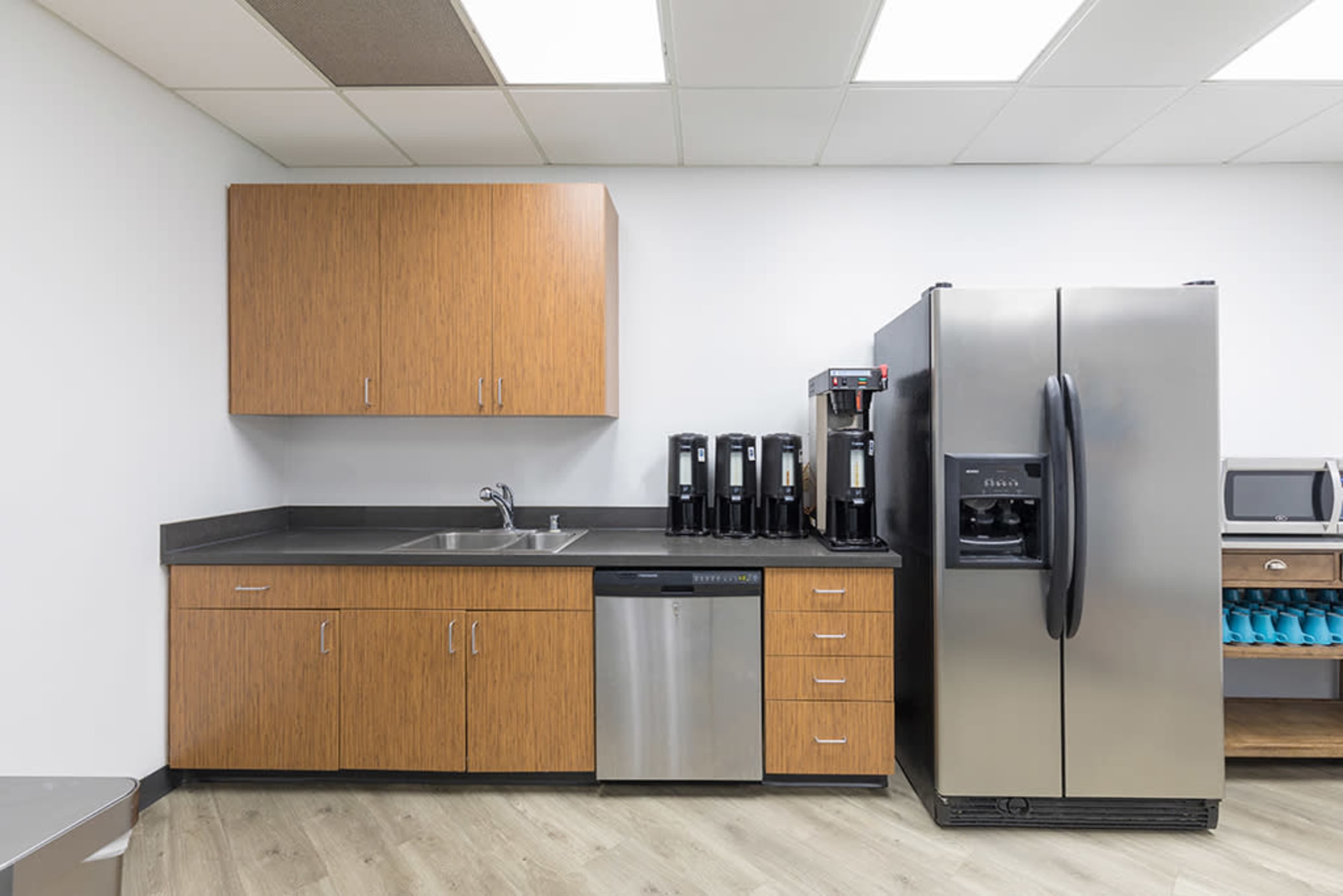 The image shows a modern kitchen with wooden cabinets, a stainless steel refrigerator, a dishwasher, and coffee machines on the counter.