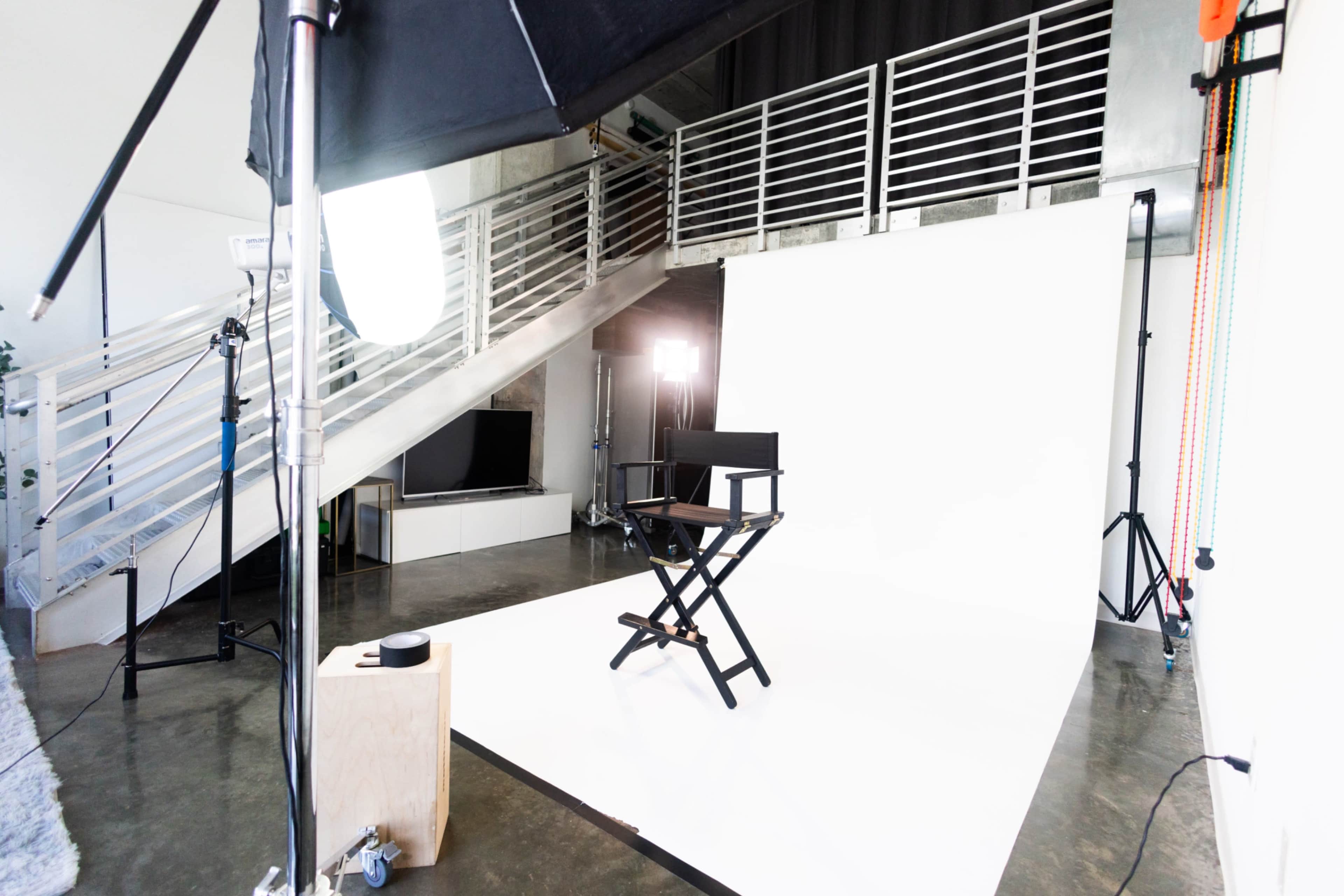 The image shows a photography studio setup featuring a blank white backdrop, a director's chair in the center, and various lighting equipment arranged around the space.