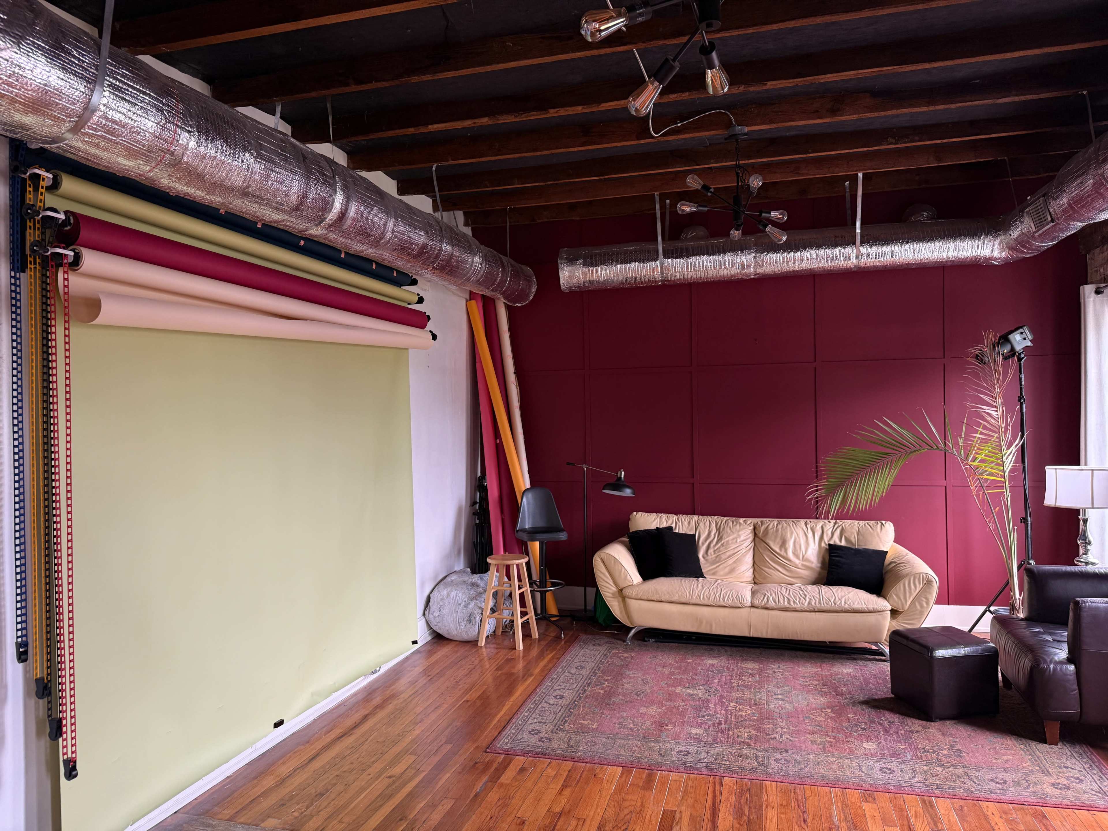 The image shows a photography studio with a green backdrop, colorful paper rolls, a beige couch, and wooden flooring.