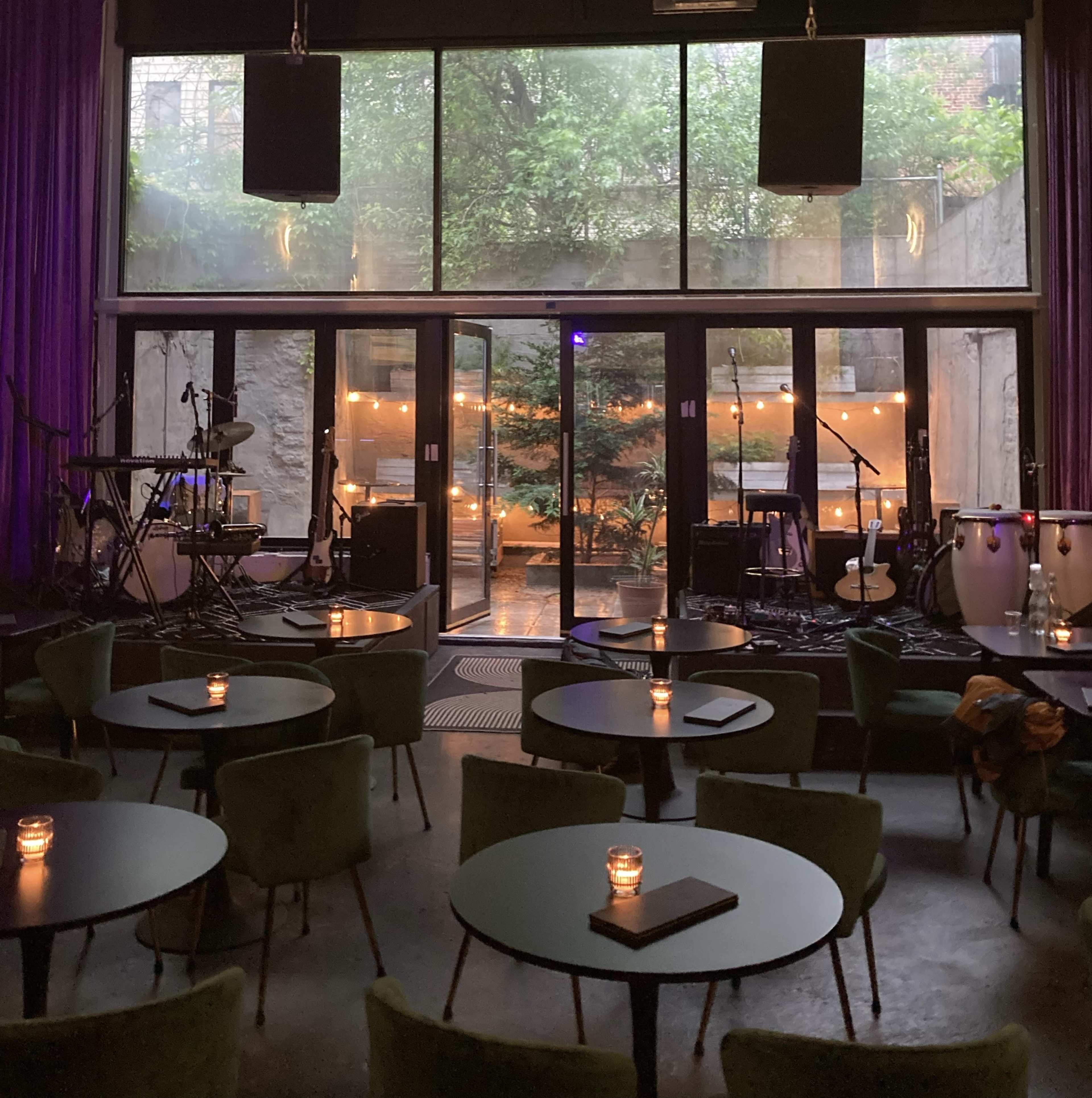 An intimate performance venue with tables and candles in the foreground, large windows revealing an outdoor space with greenery and soft lighting in the background.