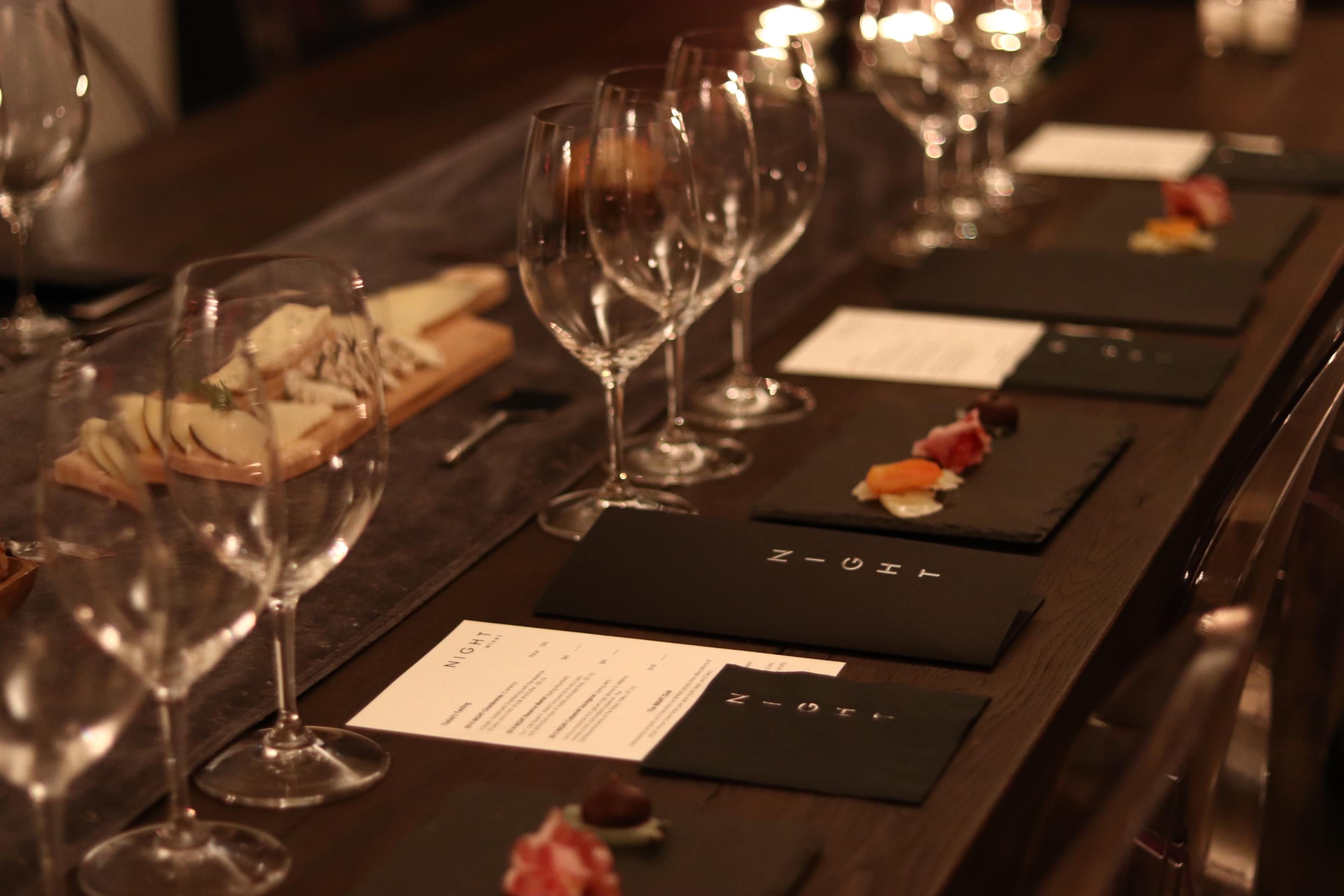 A dining table is set with wine glasses, menus, and small plates of food arranged on dark slate.