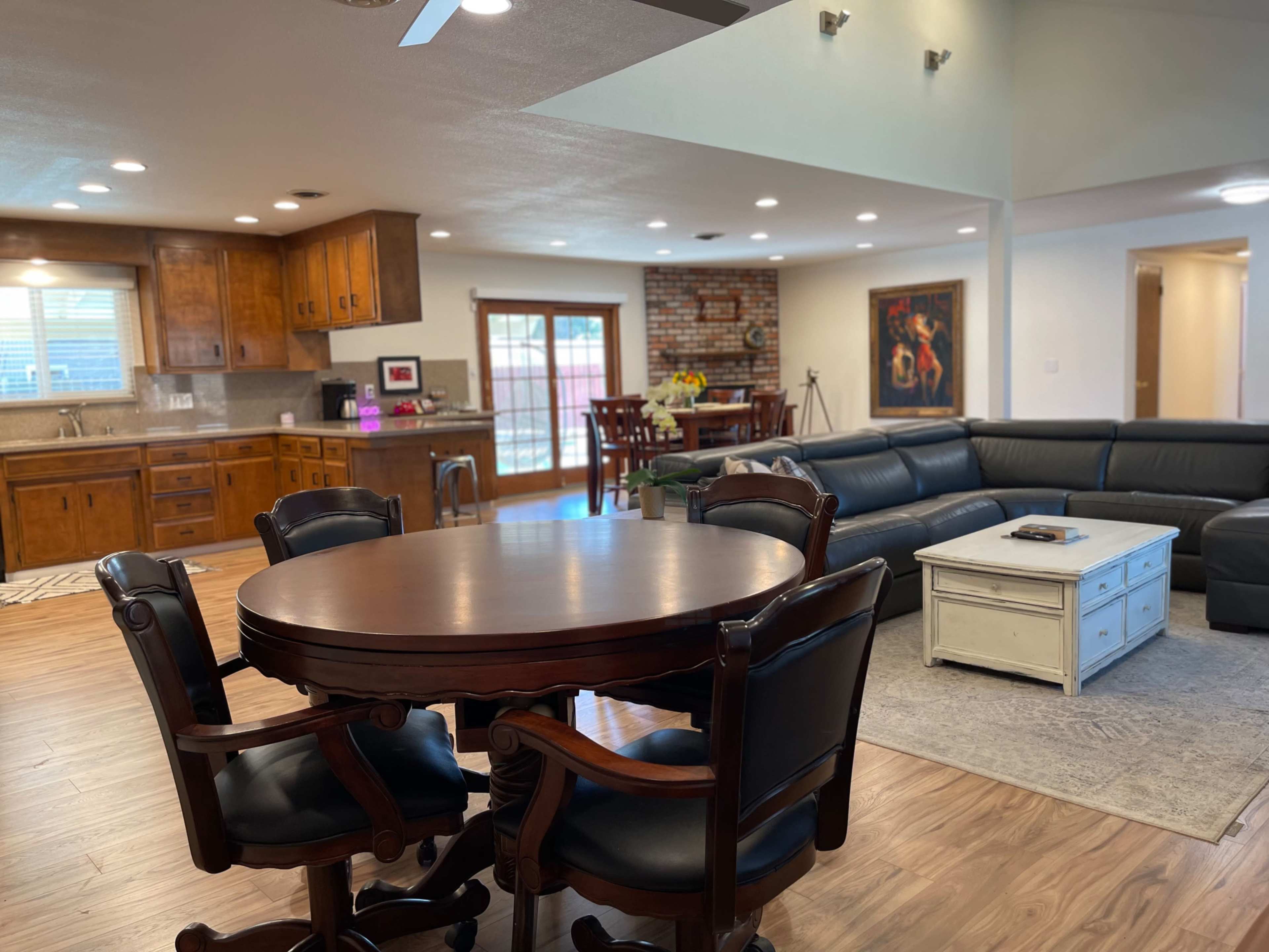 The image shows an open-concept living space with a dining table, a kitchen area with wooden cabinets, and a sitting area featuring a dark leather sectional couch.