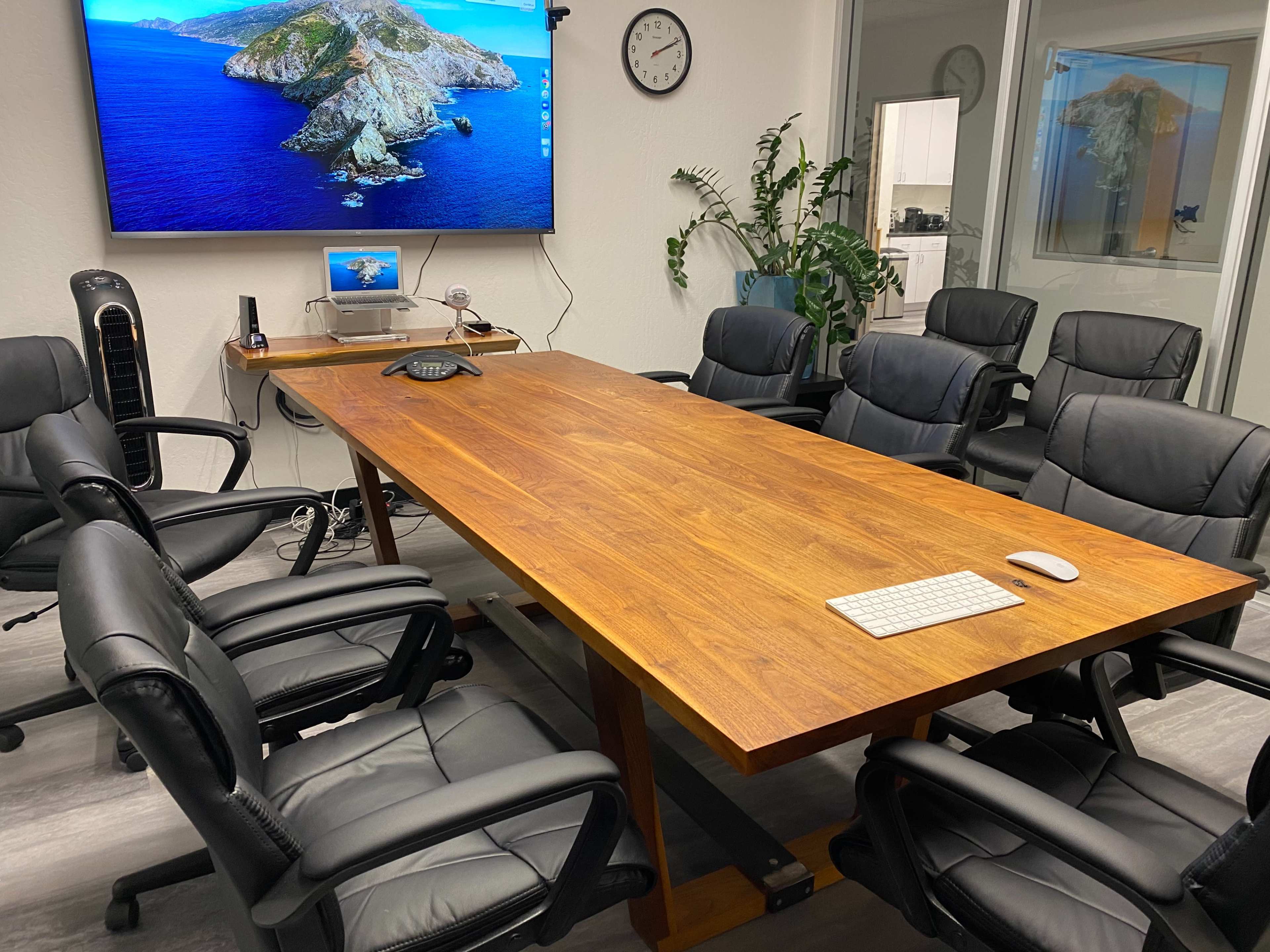 A large wooden conference table surrounded by black leather chairs is positioned in a meeting room with a wall-mounted screen displaying a coastal landscape.