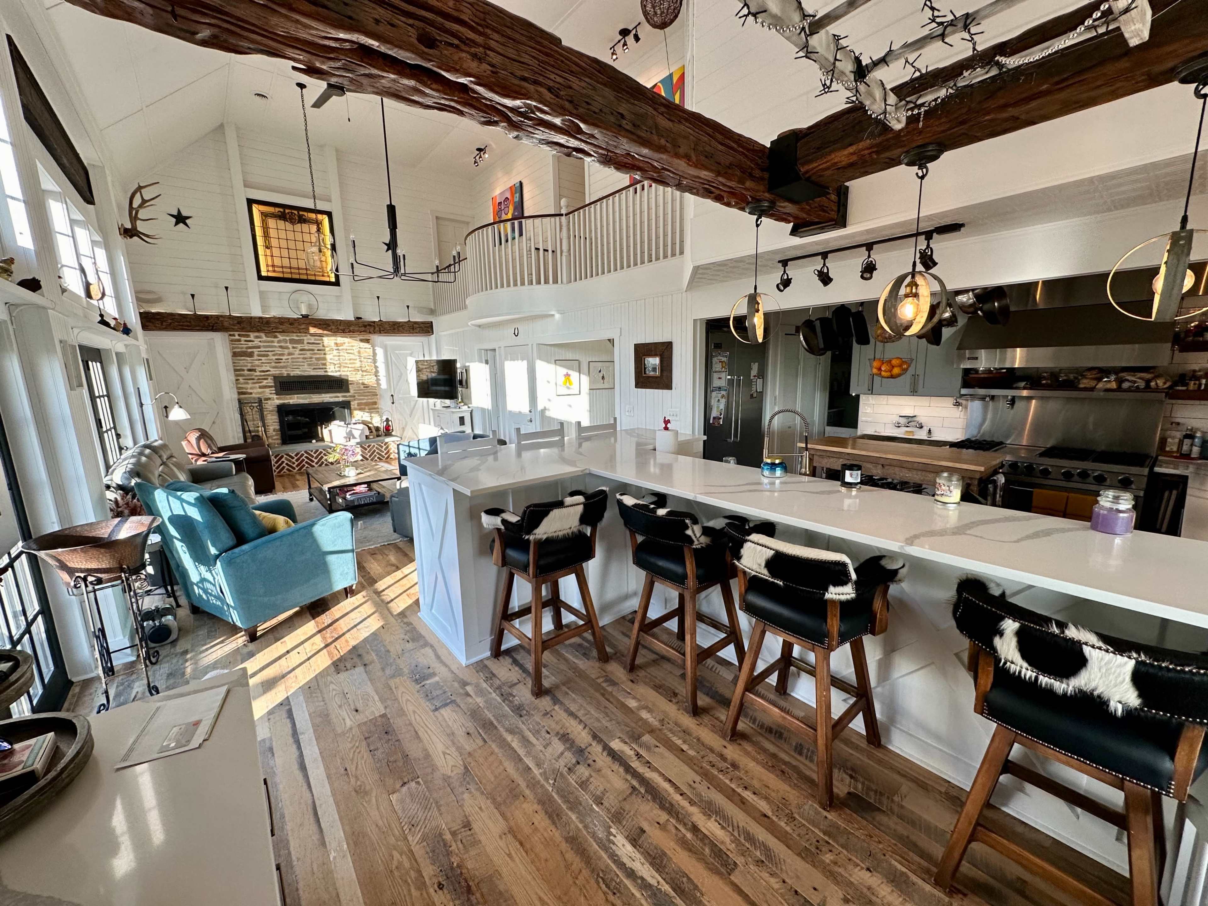 The image shows a bright, open-concept kitchen and living area with wooden beams, a large island with bar stools, and a cozy seating arrangement beside a fireplace.
