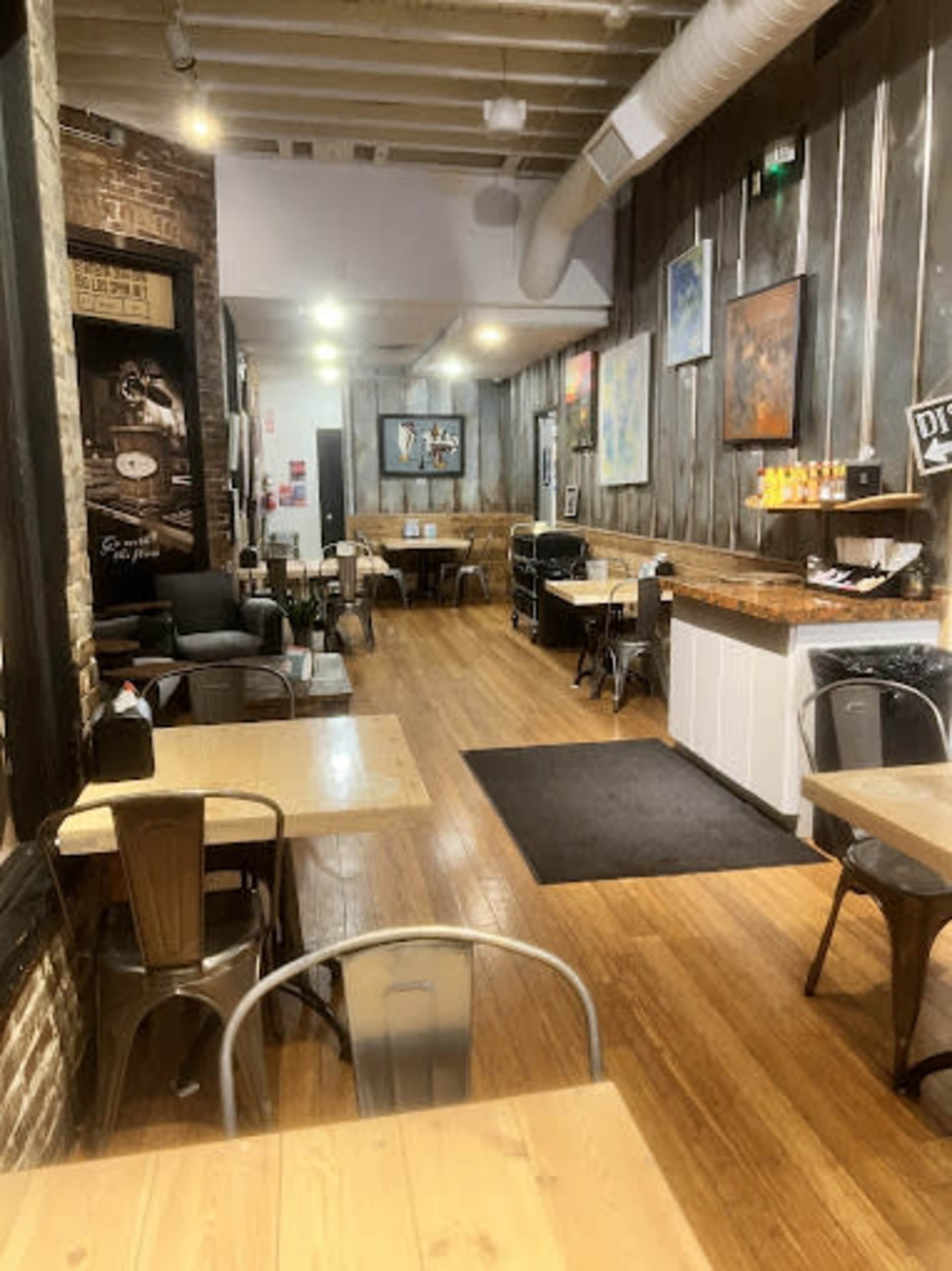 The image shows a spacious cafe with wooden floors, gray walls adorned with artwork, and various tables set for customers.