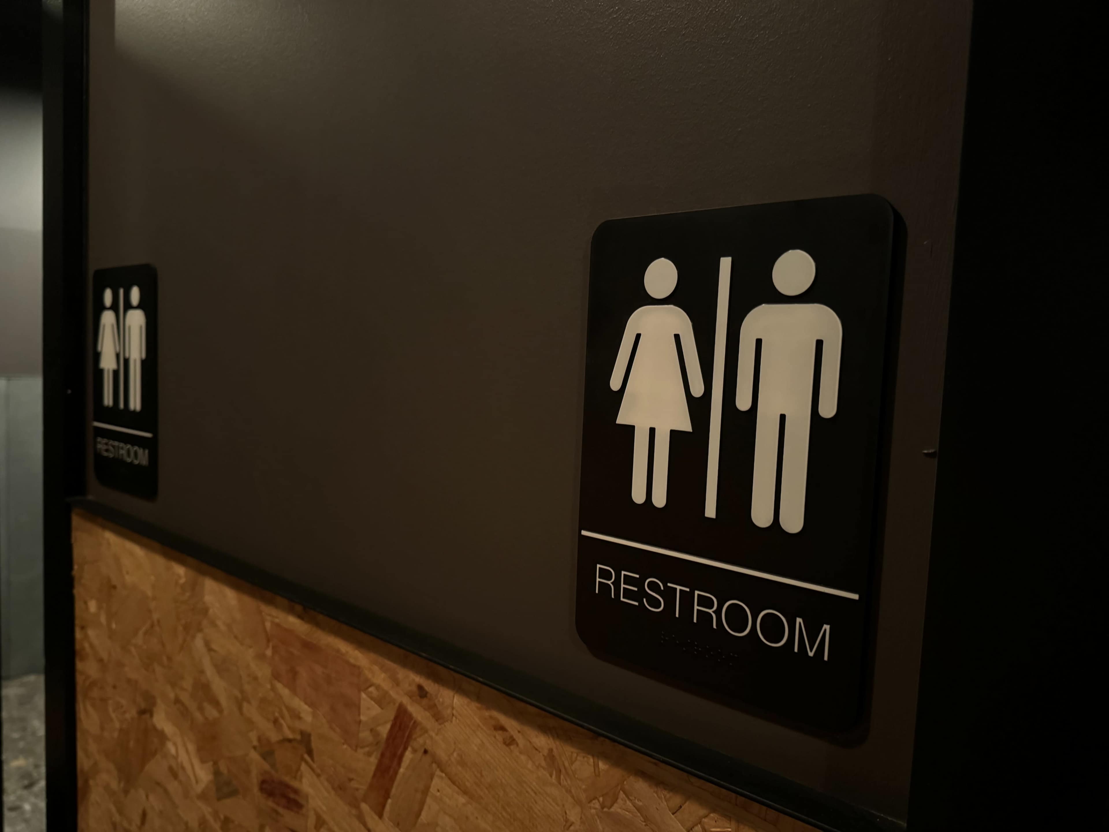 The image shows restroom signs indicating male and female facilities on a wall with an exposed wooden panel.