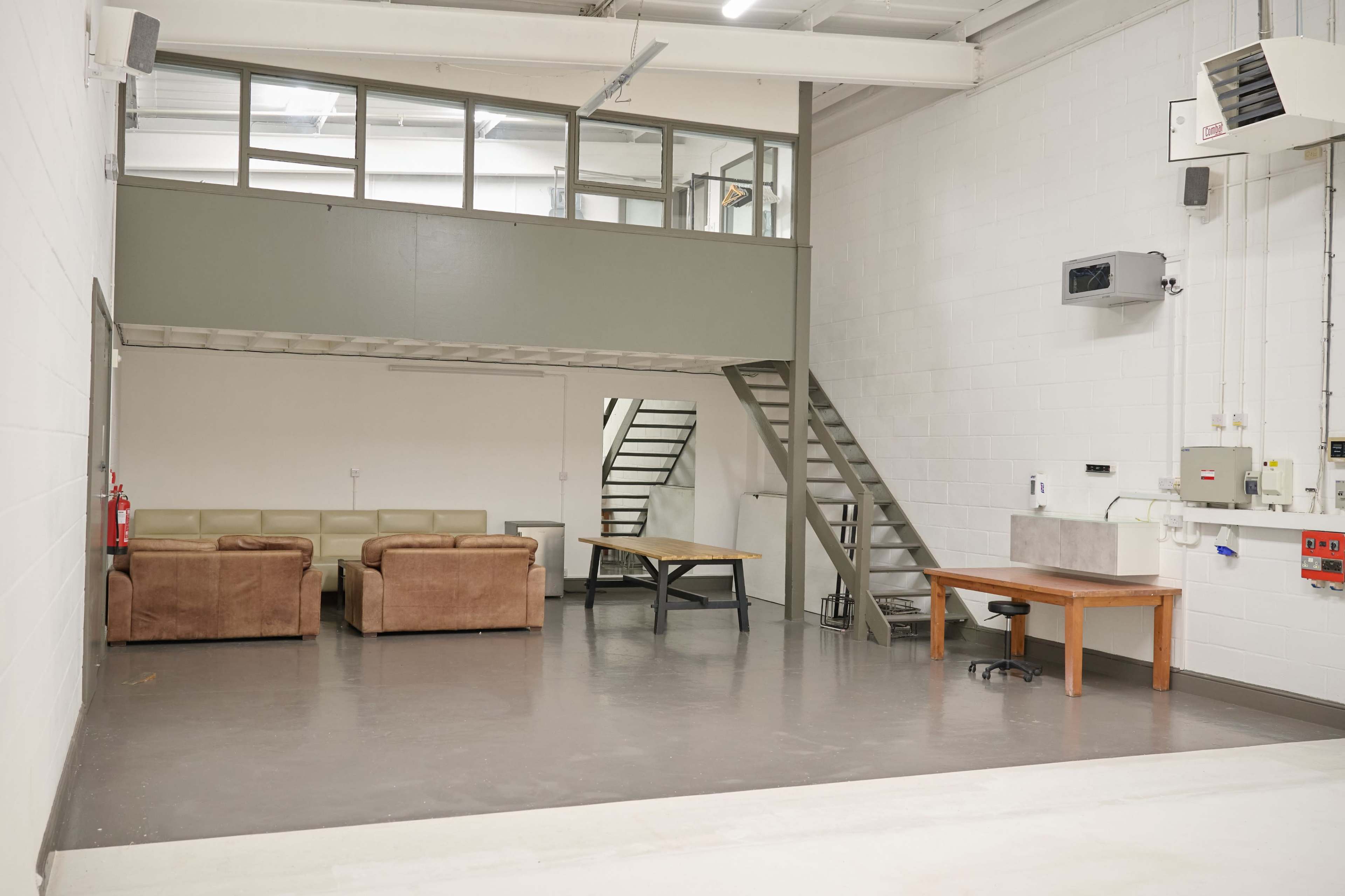 The image shows a spacious, industrial-style room featuring two brown couches, a staircase, and several wooden tables against plain white walls.