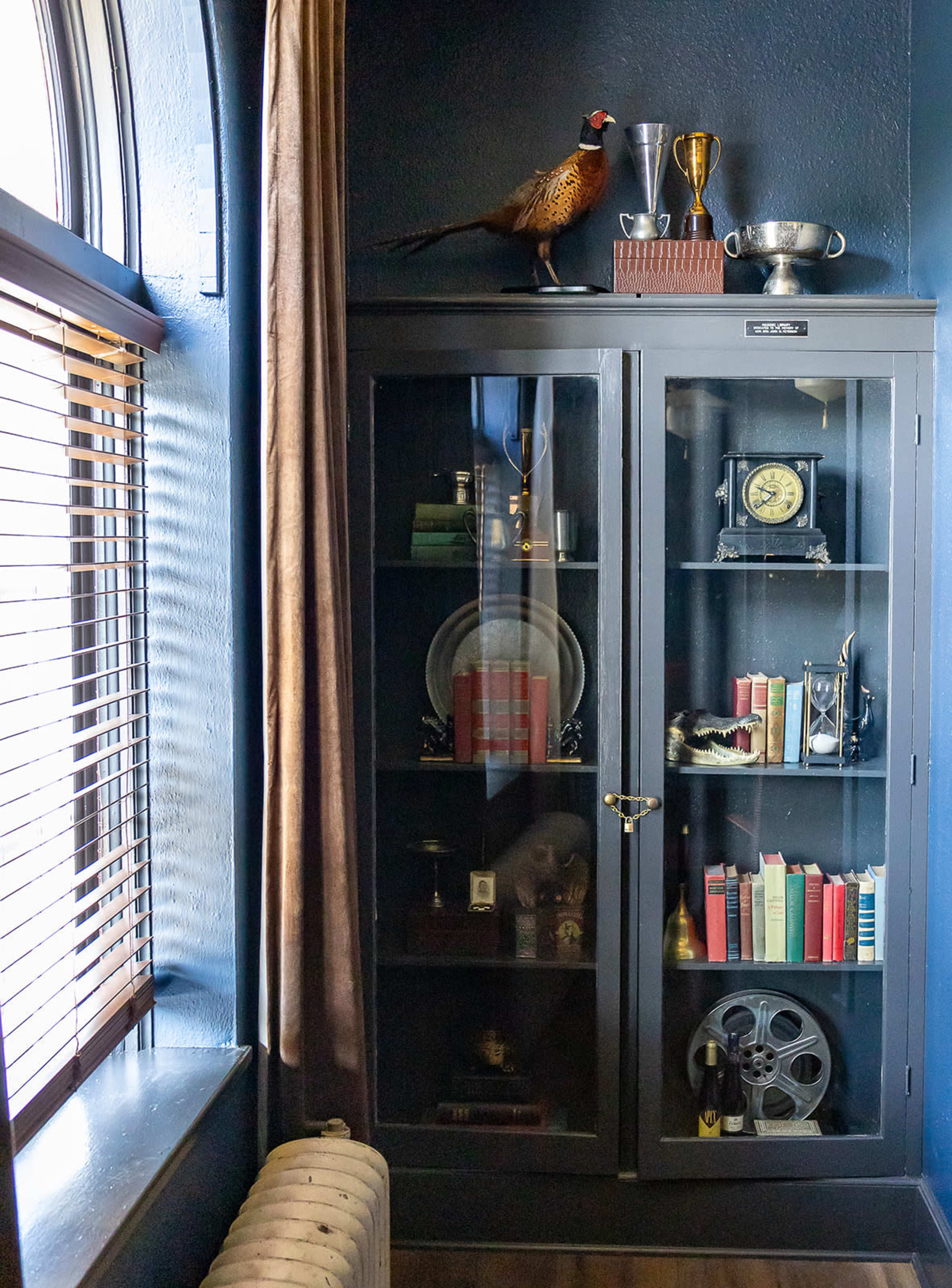 A glass-fronted cabinet displays various vintage items, including books, a clock, and trophies, in a corner of a room with a window featuring blinds.