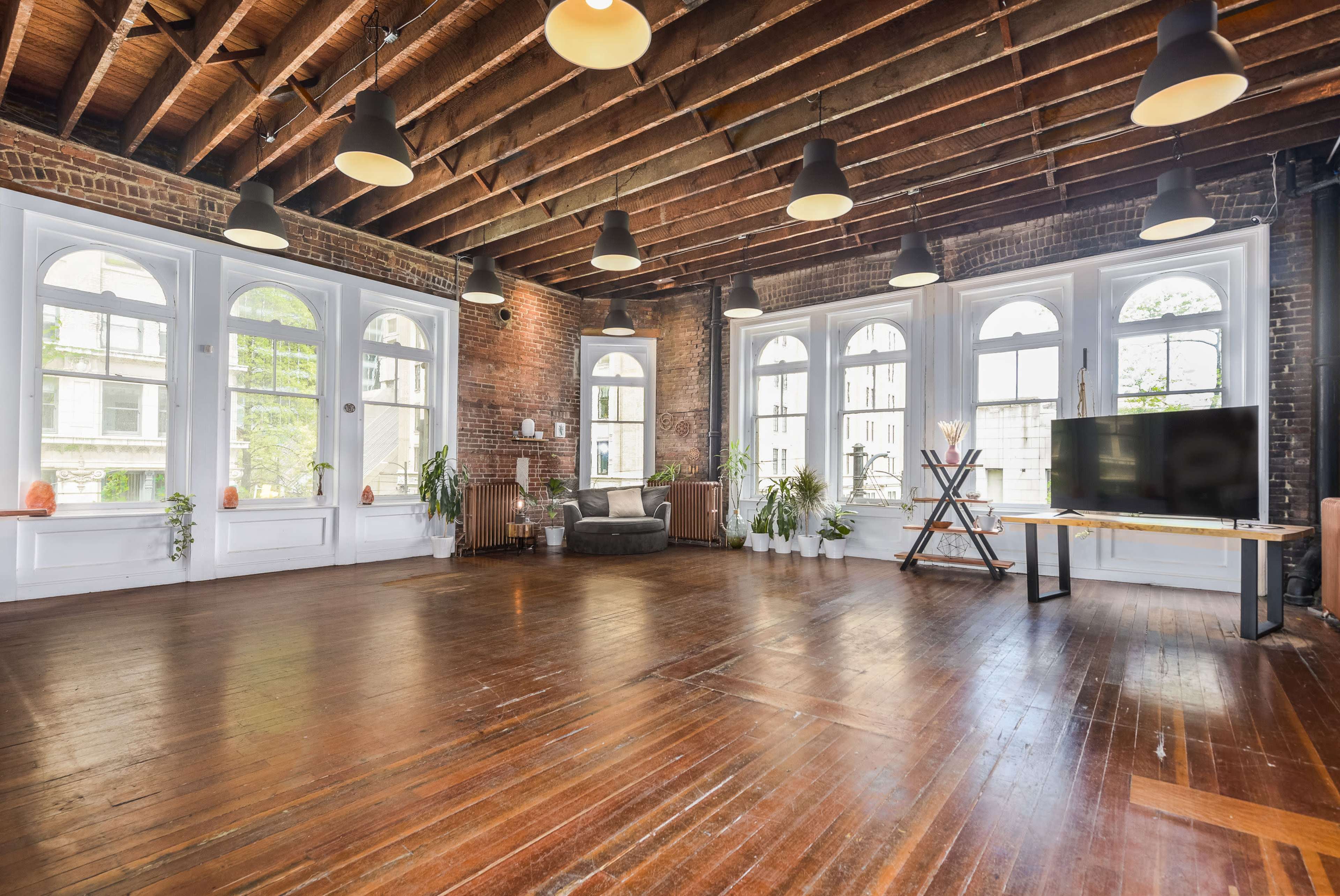 A spacious interior room with exposed wooden beams, large windows, and a mix of modern and rustic decor, featuring hardwood floors and potted plants.