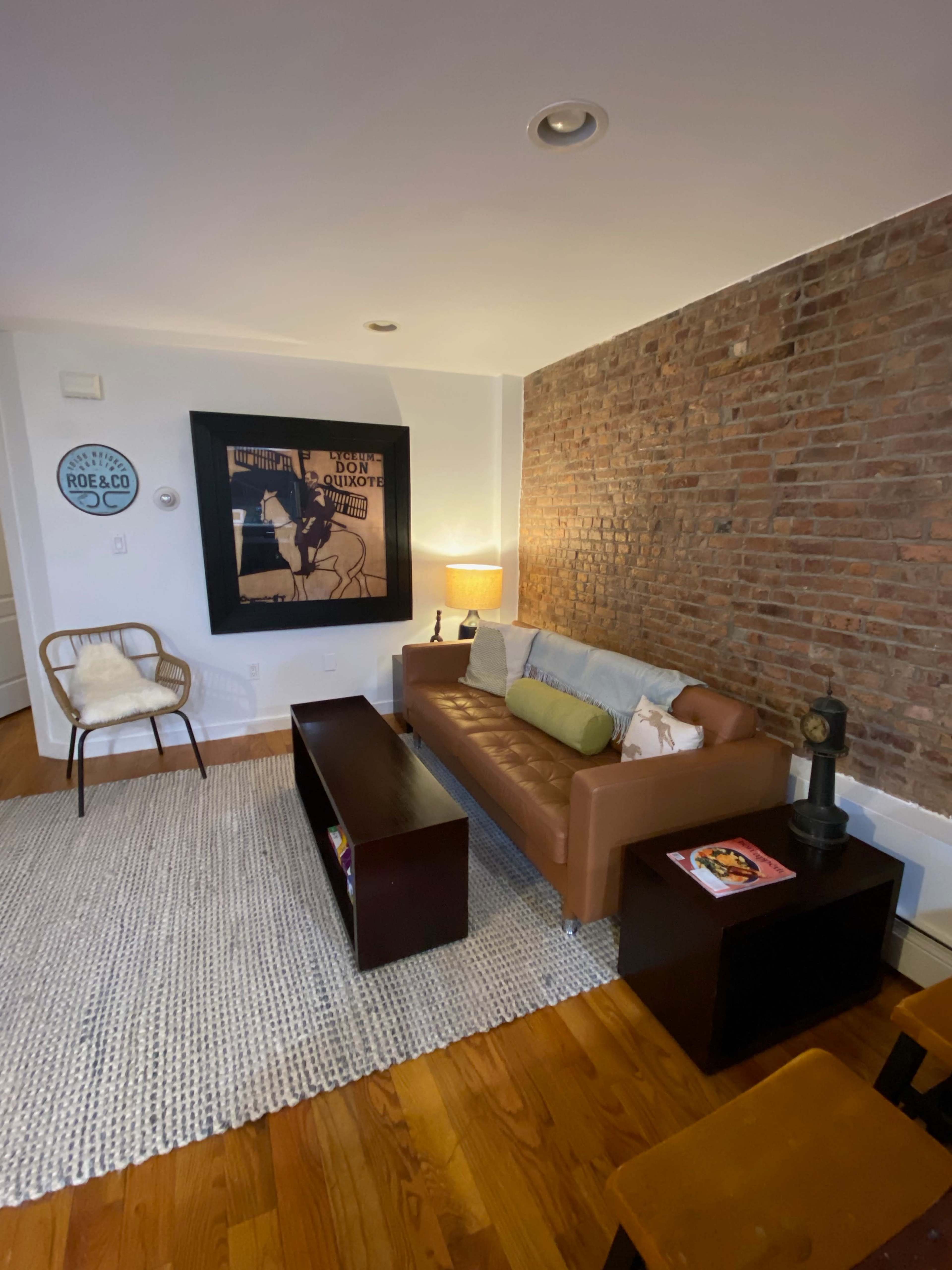 A cozy living room featuring a brown leather sofa, a wooden coffee table, a chair, and a textured rug against a brick wall.