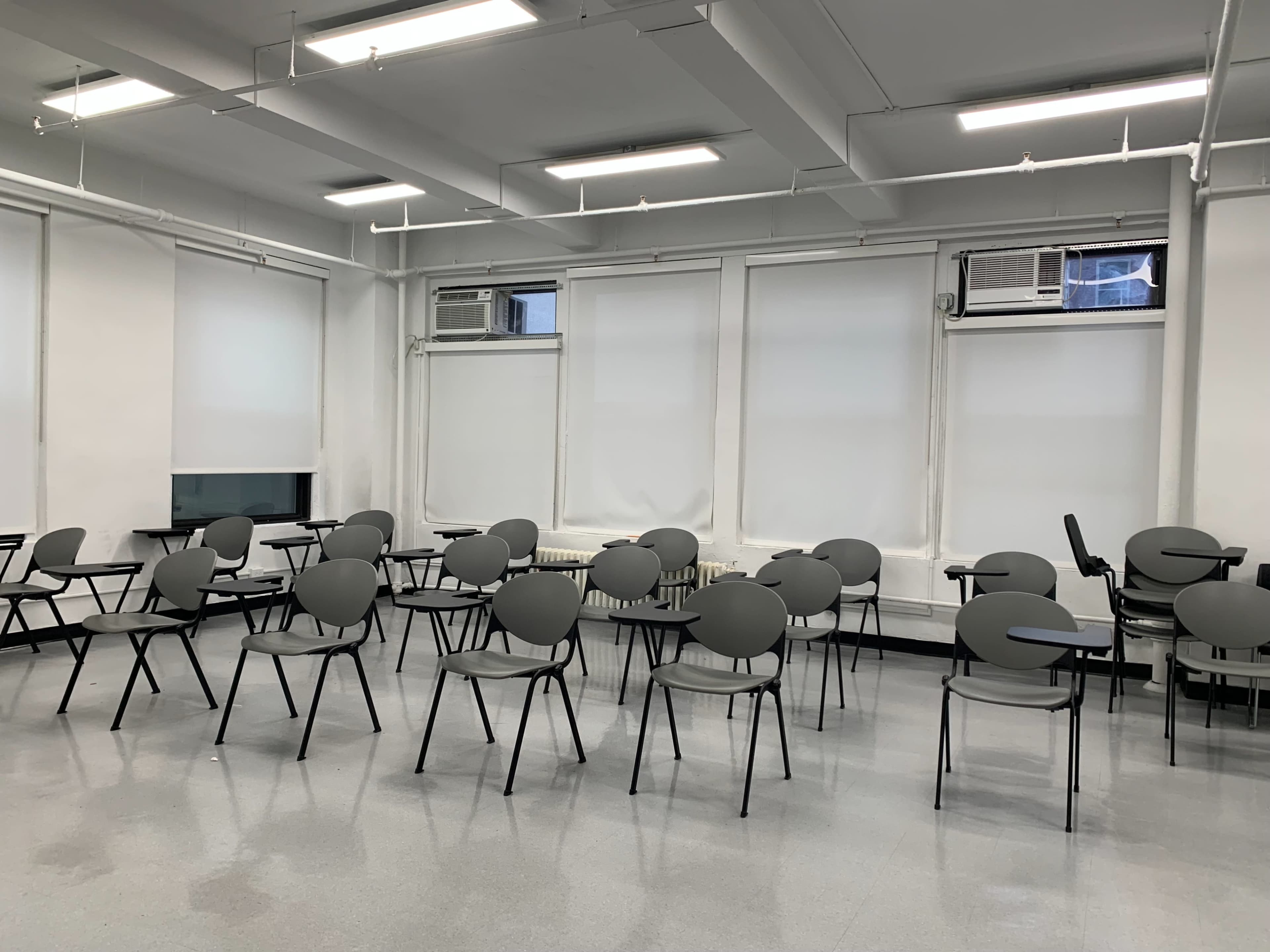 A classroom features several grey chairs and desks arranged in rows, with windows covered by blinds and air conditioning units visible.