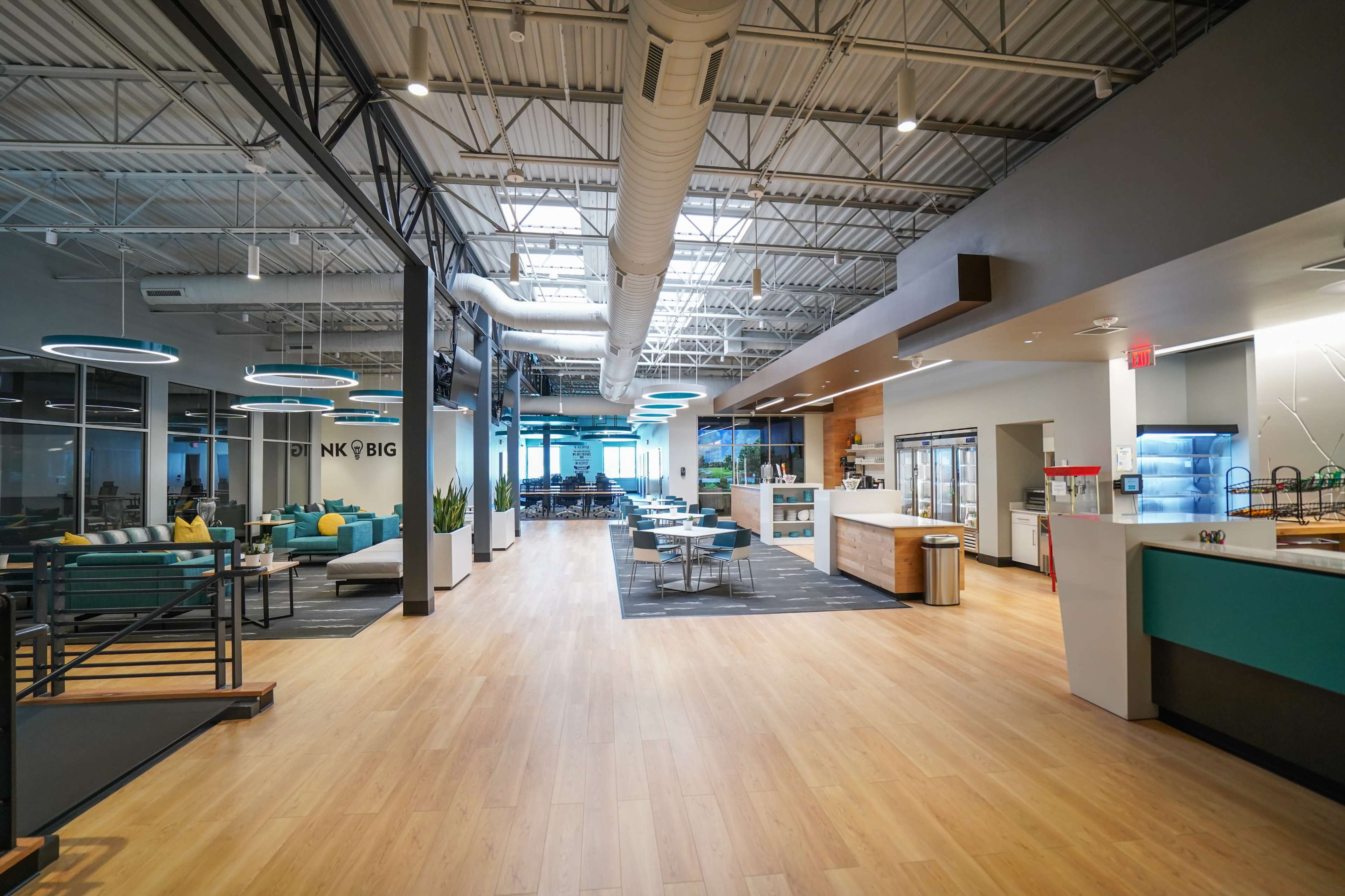 A spacious, modern office interior features a café area with seating, a kitchen counter, and large windows allowing natural light to enter.