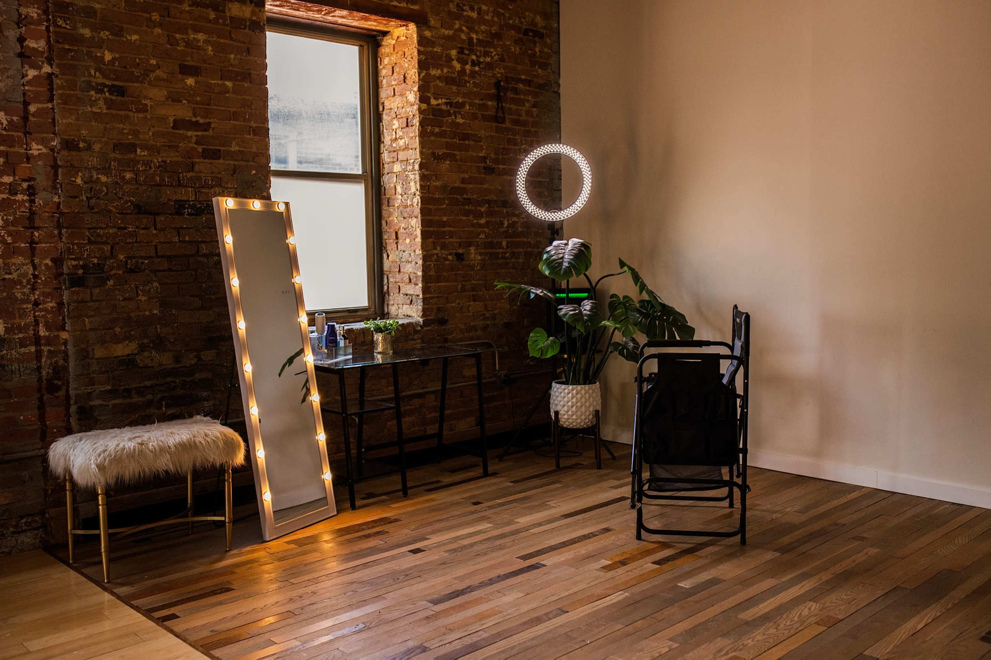 The image shows a well-lit room with a brick wall, featuring a large mirror with lights, a table with plants, a round mirror, and a chair.