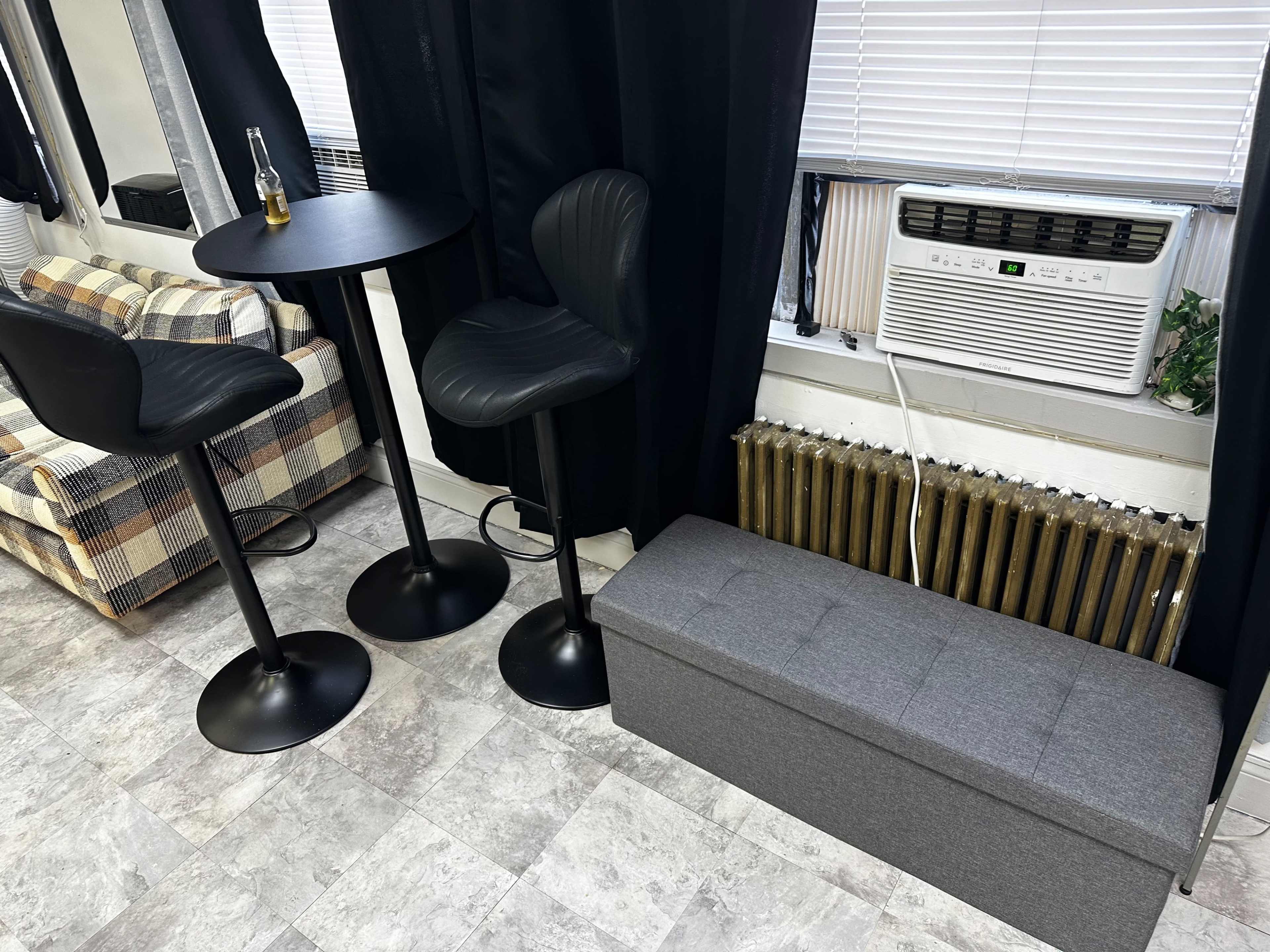 The image shows a small interior space featuring two tall black bar stools at a round table, an air conditioning unit by the window, and a gray bench near a radiator.