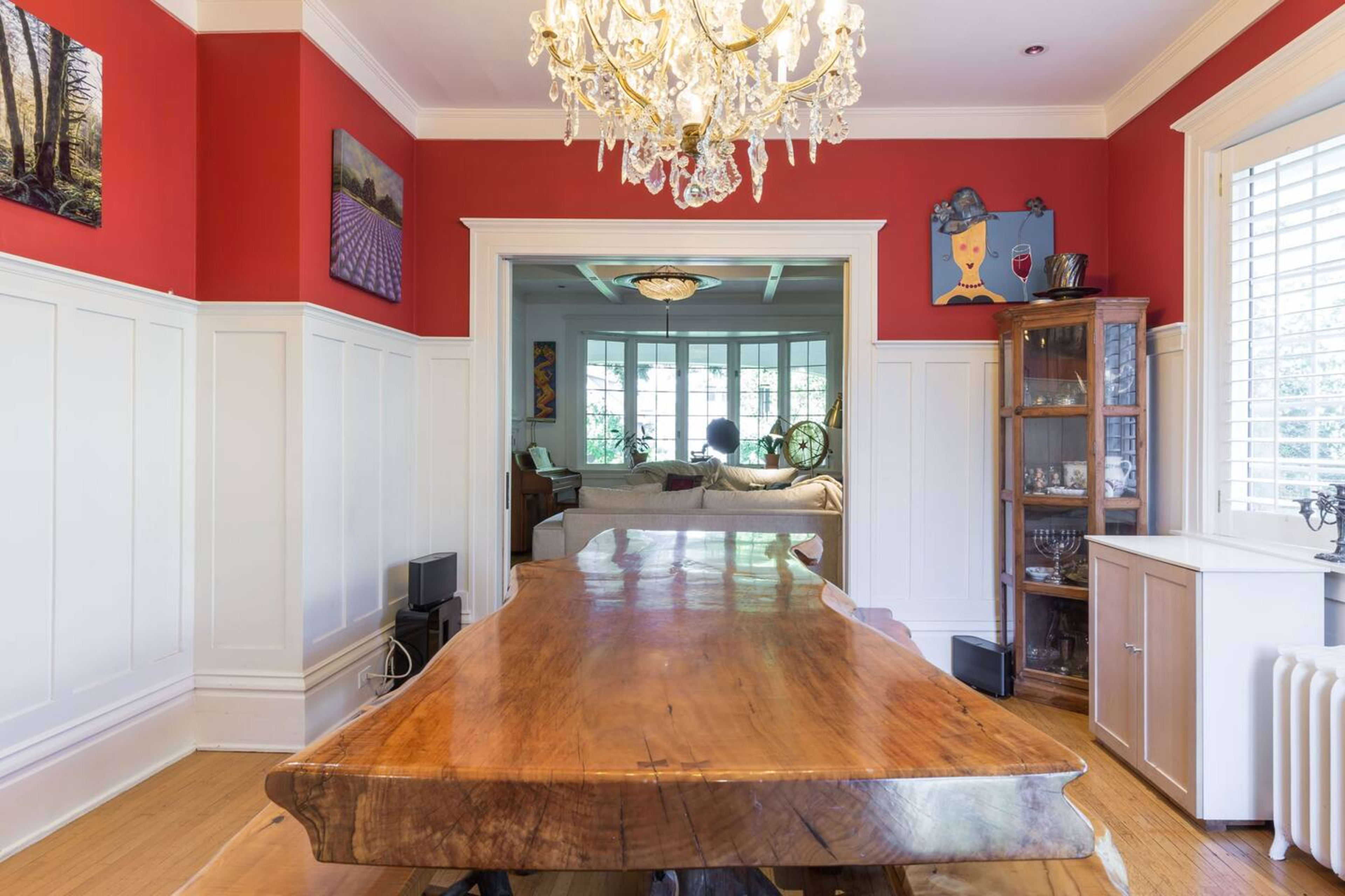 A long wooden dining table is the focal point of a room with red walls, framed artwork, and a chandelier above.